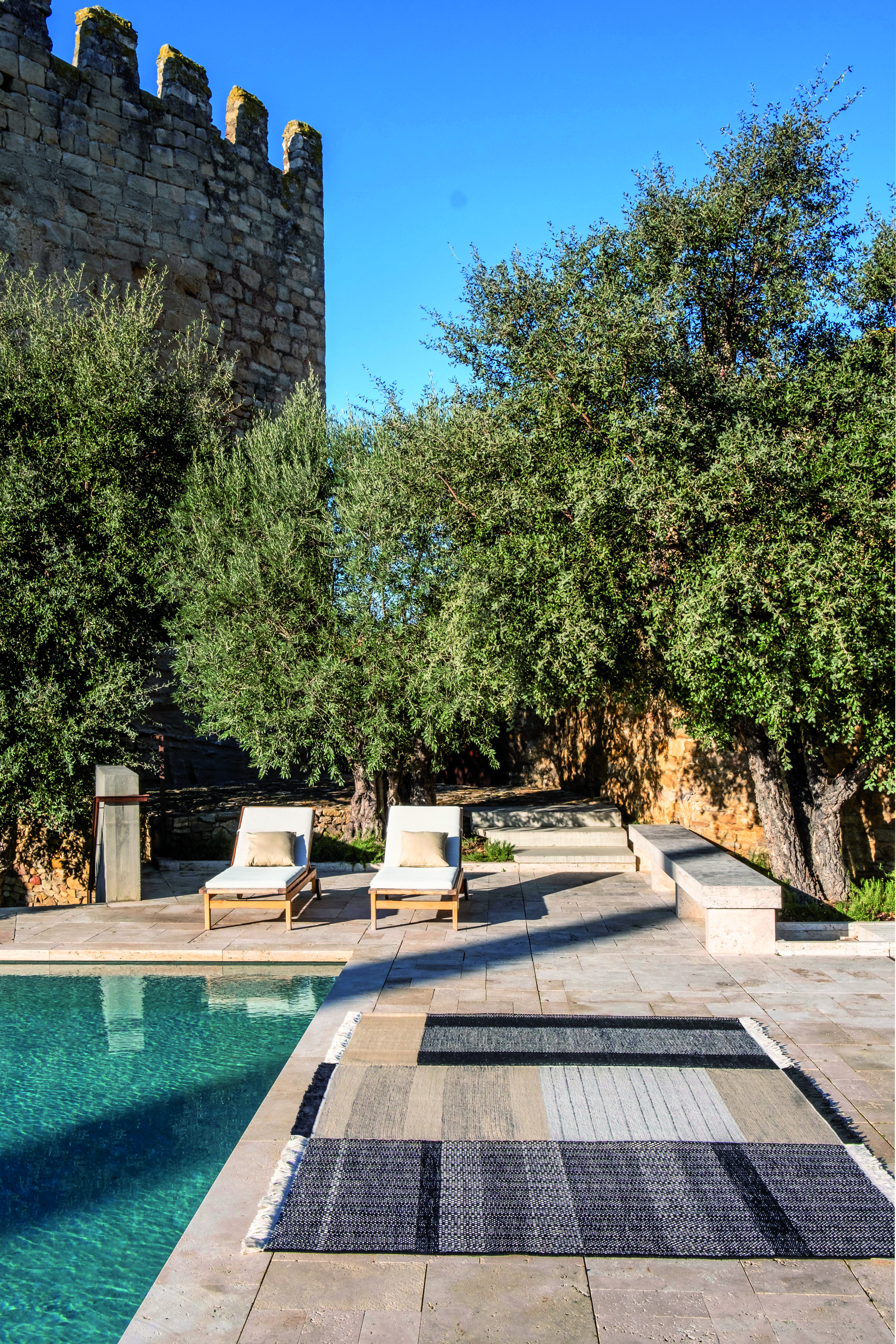 Teppich am Pool auf Natursteinboden unter Bäumen mit Liegestühlen  