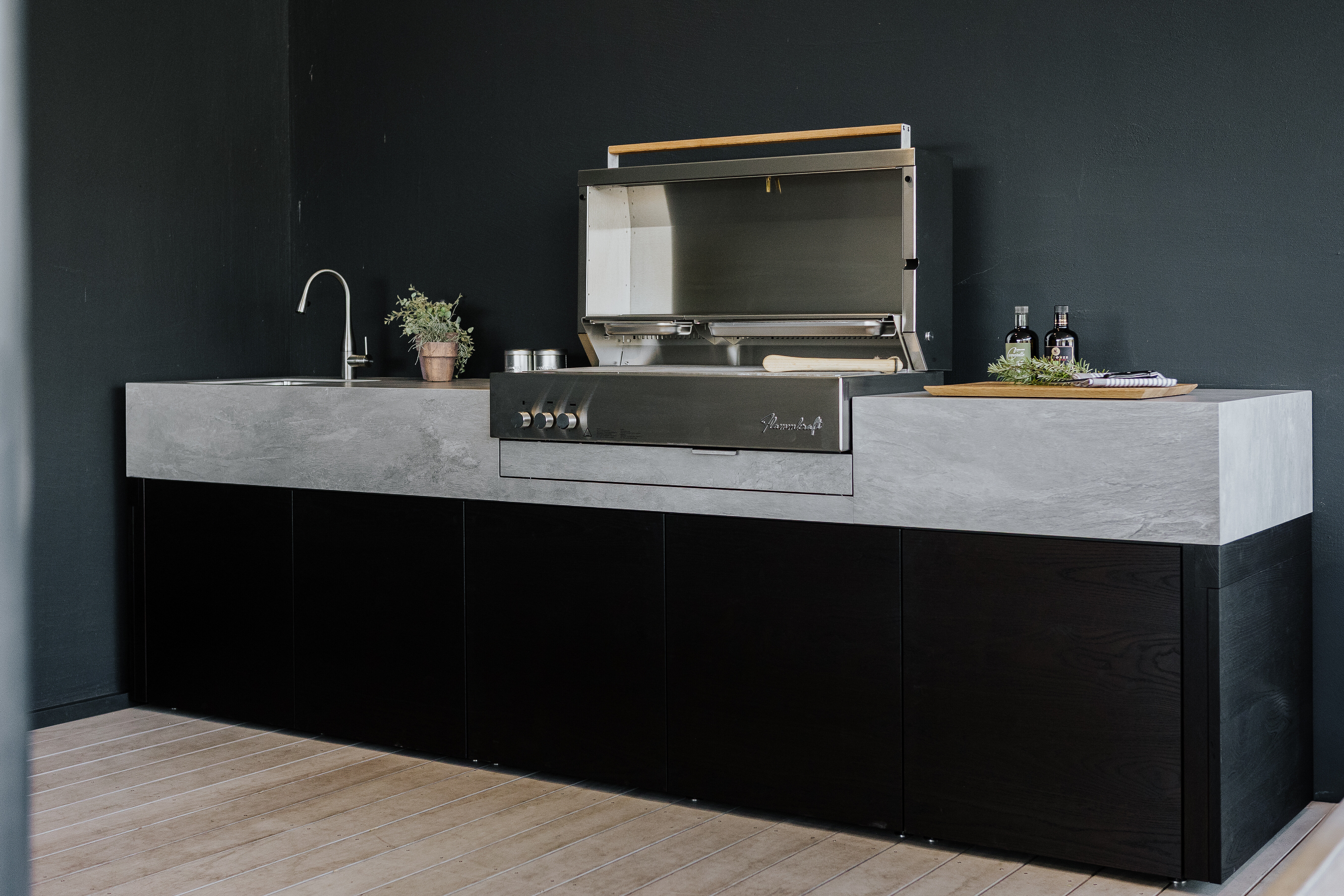 Outdoor-Küche mit dunklen Fronten und heller Arbeitsplatte, eingebautem Grill und Spülbecken. 