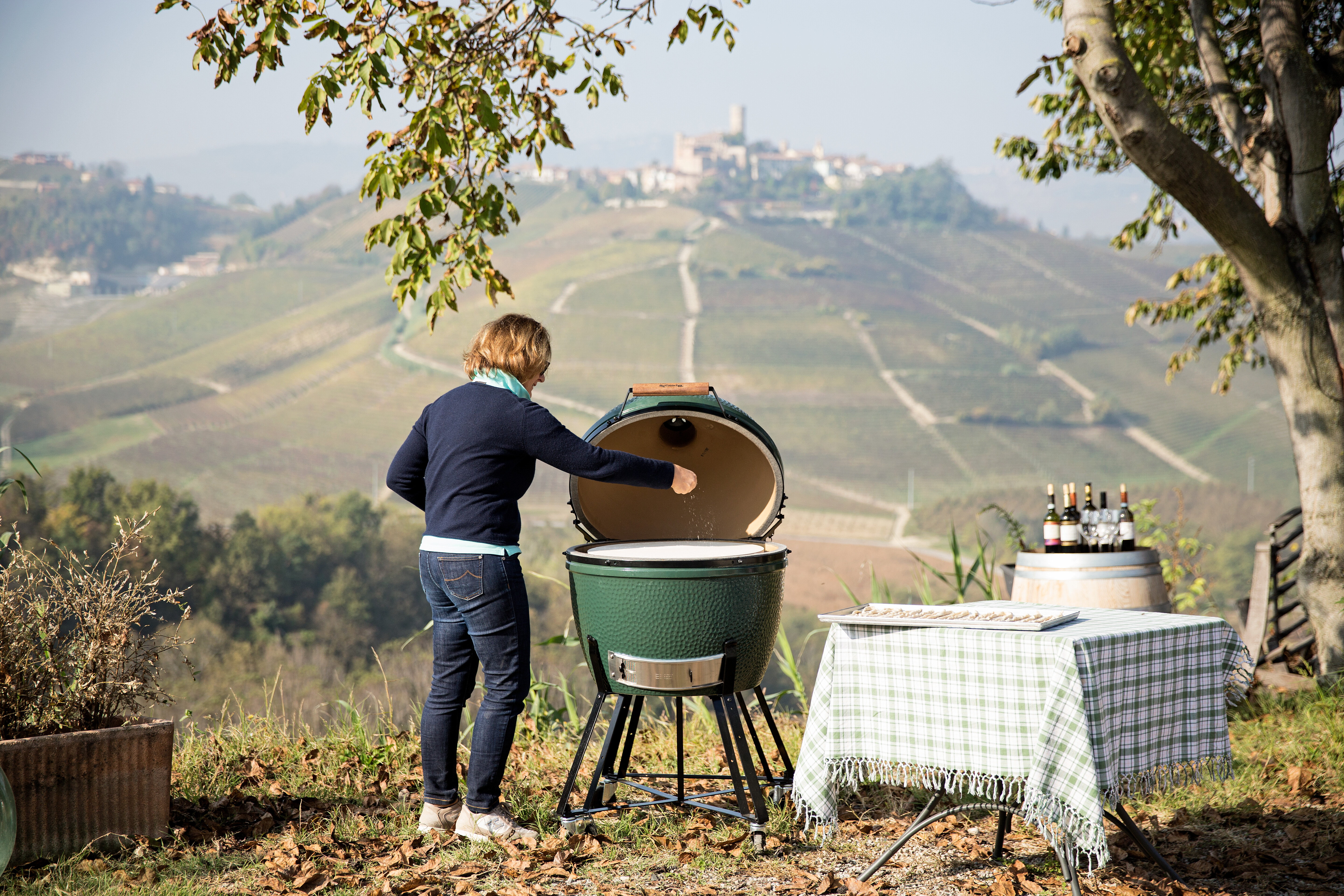 Person bedient großes Big Green Egg Modell mit Ausblick auf Weinberge und Hügellandschaft  