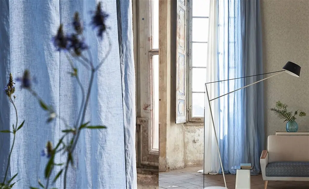 Halbtransparente Vorhänge in Hellblau vor großen Fenstern mit floraler Dekoration im Vordergrund  