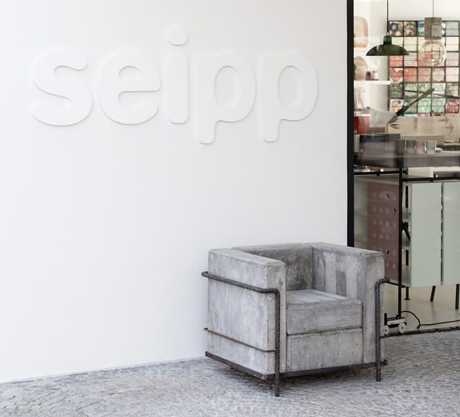 Nahaufnahme des Beton-LC2 Sessels von Stefan Zwicky vor dem Seipp Eingang in Waldshut mit Sicht auf weißen Fassadenschriftzug und Showroom-Einblick.