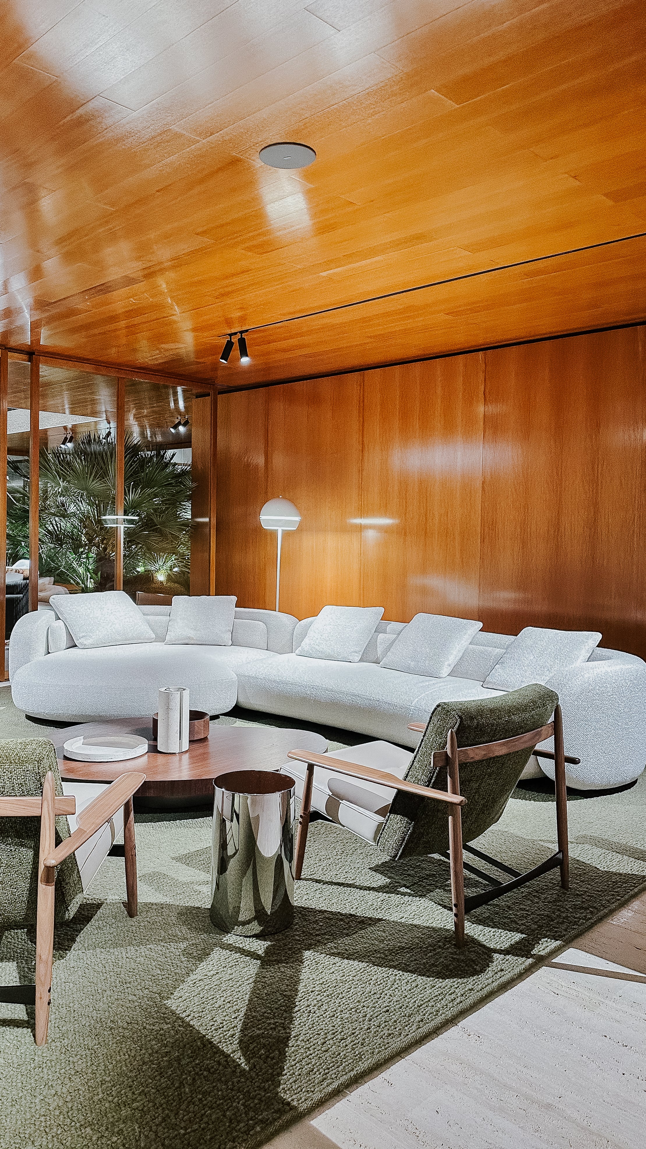 Wohnszene auf der Il Salone del Mobile 2025 mit weißem Rundsofa, zwei Sesseln mit Holzgestell, runder Tischgruppe und grünem Teppich. Raum mit Holzdecke, Wandverkleidung und großem Fensterbereich.