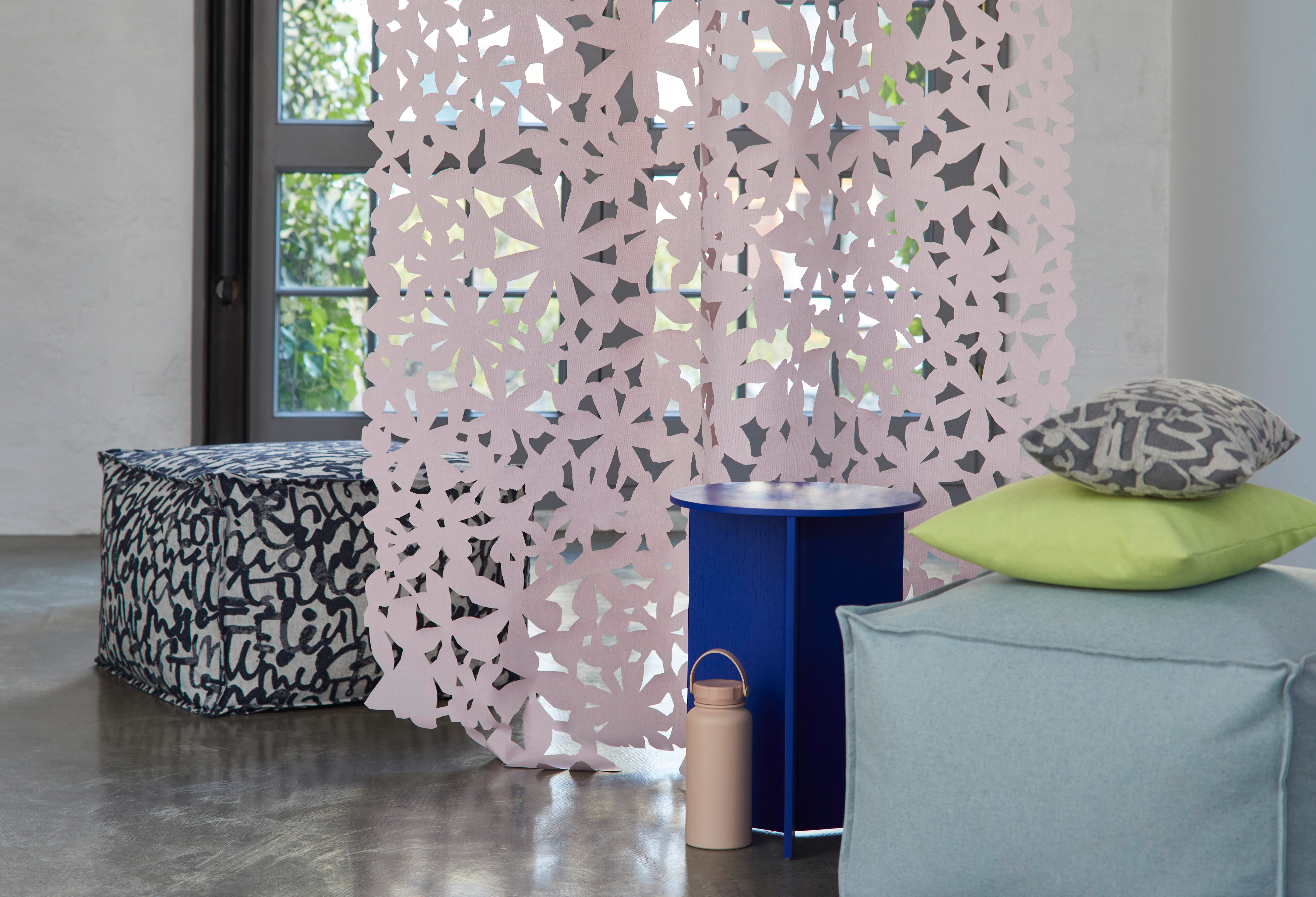 Raum mit filigranem Raumteiler in Rosa, umgeben von Poufs, Kissen und farbigen Beistelltischen  