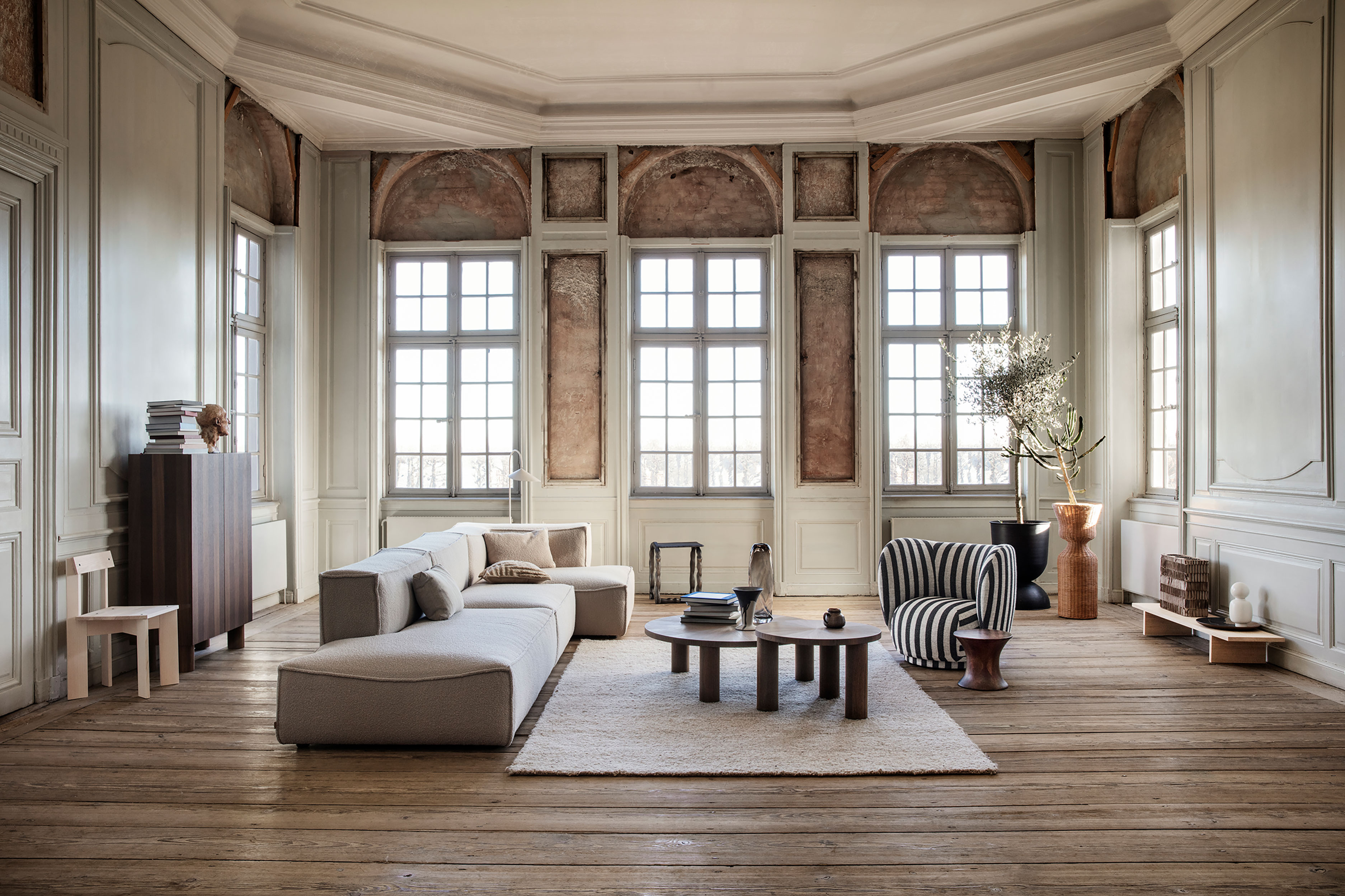 Heller Wohnraum mit Sofa, Sessel, Couchtischen und Holzfußboden vor großen, historischen Fenstern