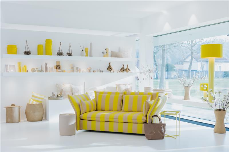 Ausstellungsansicht bei Seipp Wohnen Tiengen mit gelb gestreiftem Sofa, farblich abgestimmten Accessoires und großem Fenster mit Blick ins Freie. Helle, harmonisch komponierte Raumstimmung.
