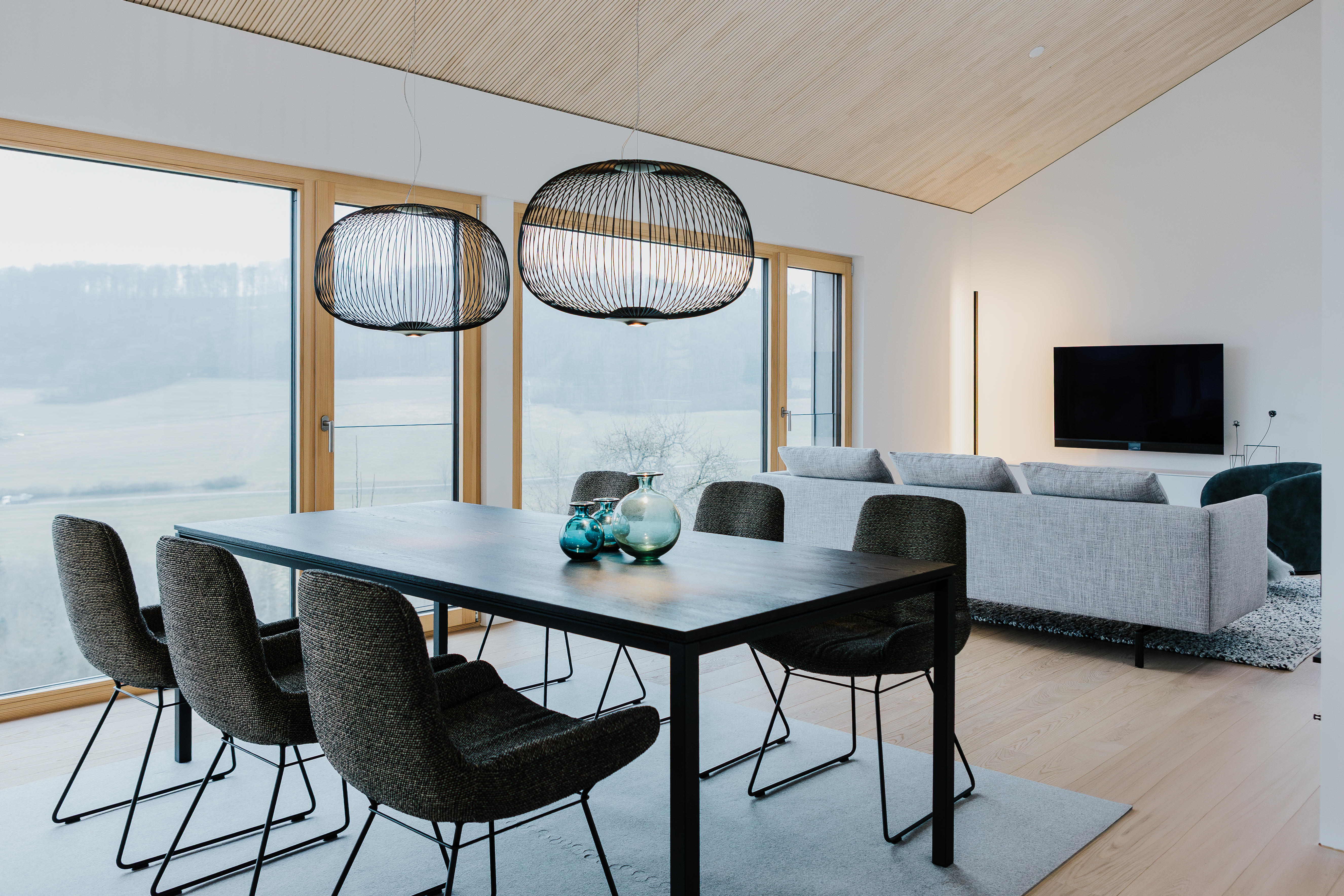 Essbereich mit dunklem Tisch und filigranen Hängelampen, dahinter Sofa und große Fenster mit Weitblick.  