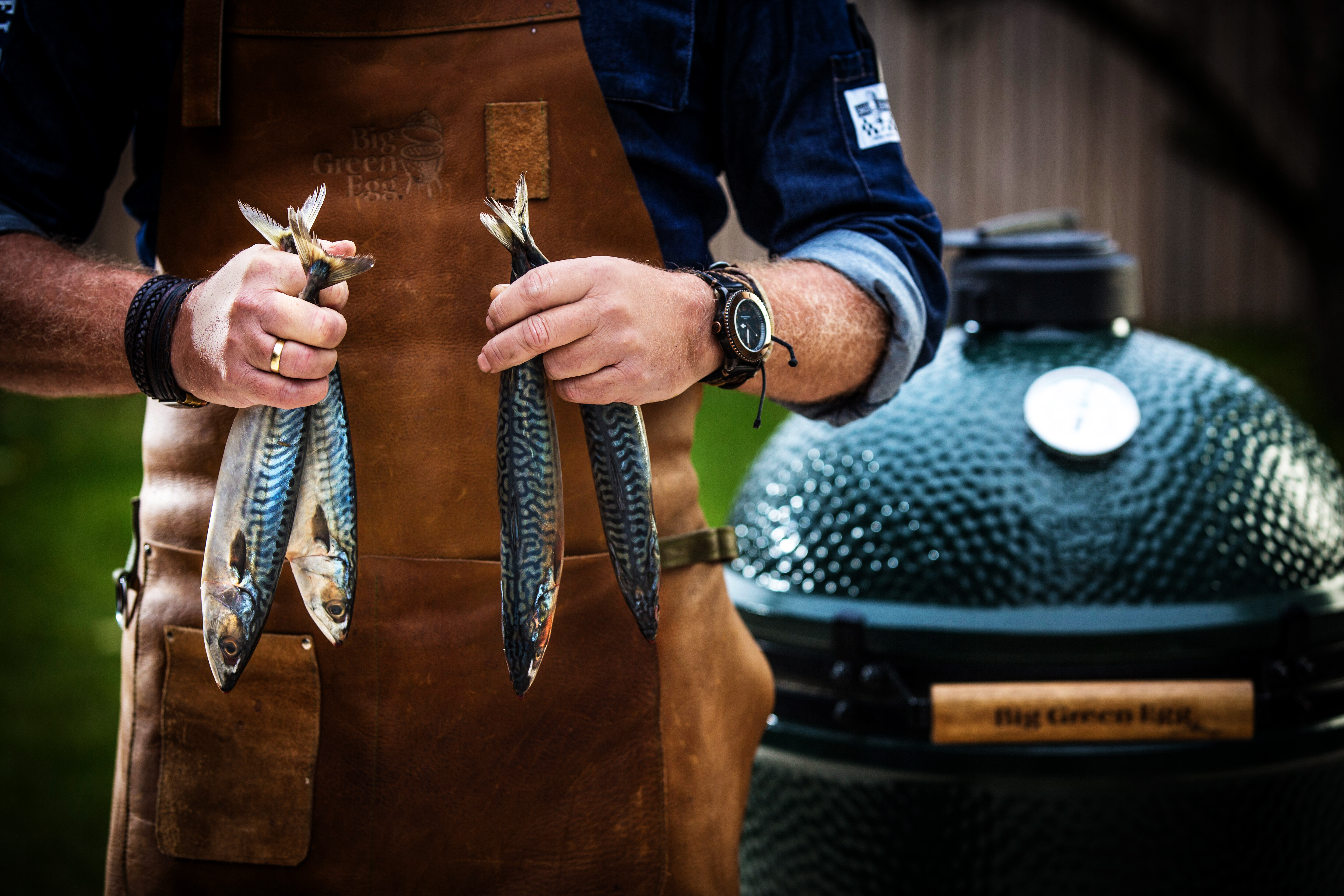 Person mit Lederschürze hält frischen Fisch, im Hintergrund ein Big Green Egg Grillgerät  