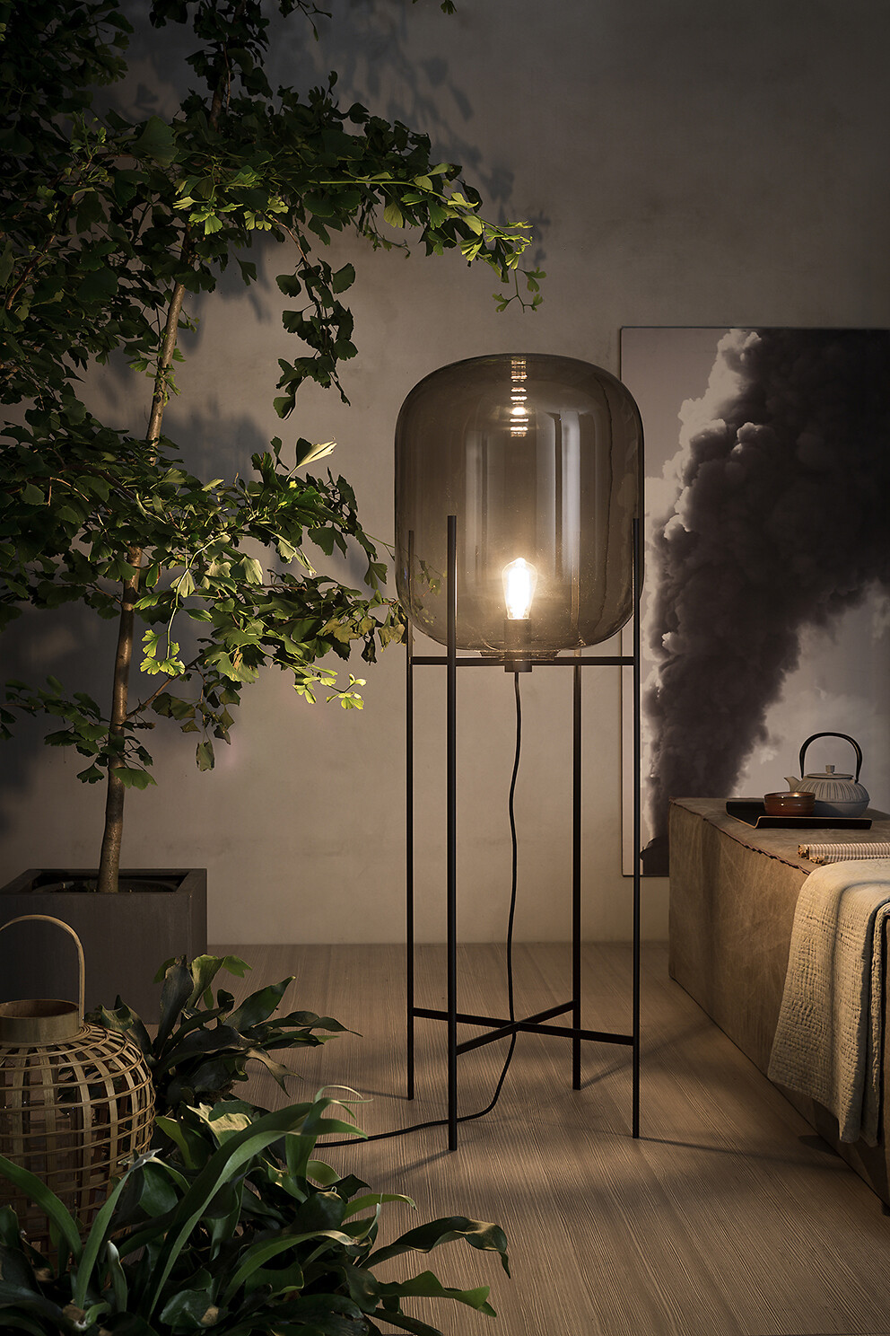 Stehleuchte mit rauchfarbenem Glaskörper neben Bett und großem Pflanzenarrangement