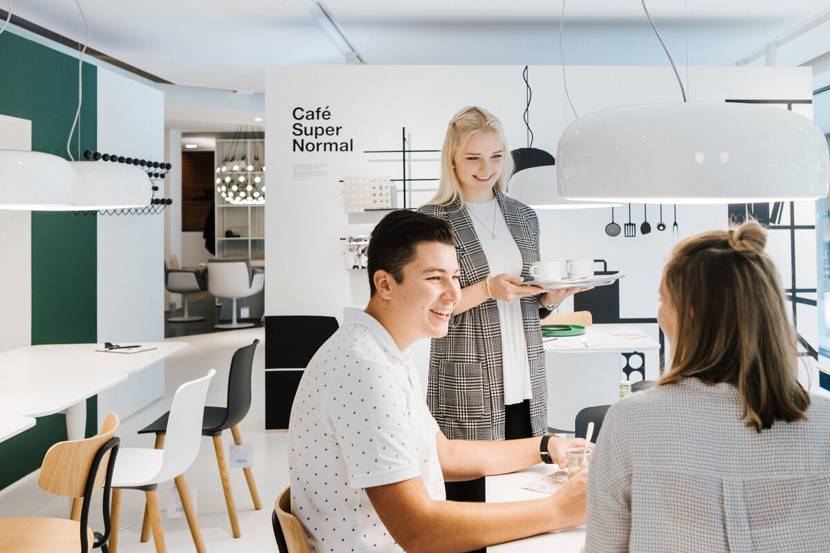 Zwei Personen im Gespräch am Tisch im Café Super Normal, dritte Person im Stehen mit Geschirr in der Hand.