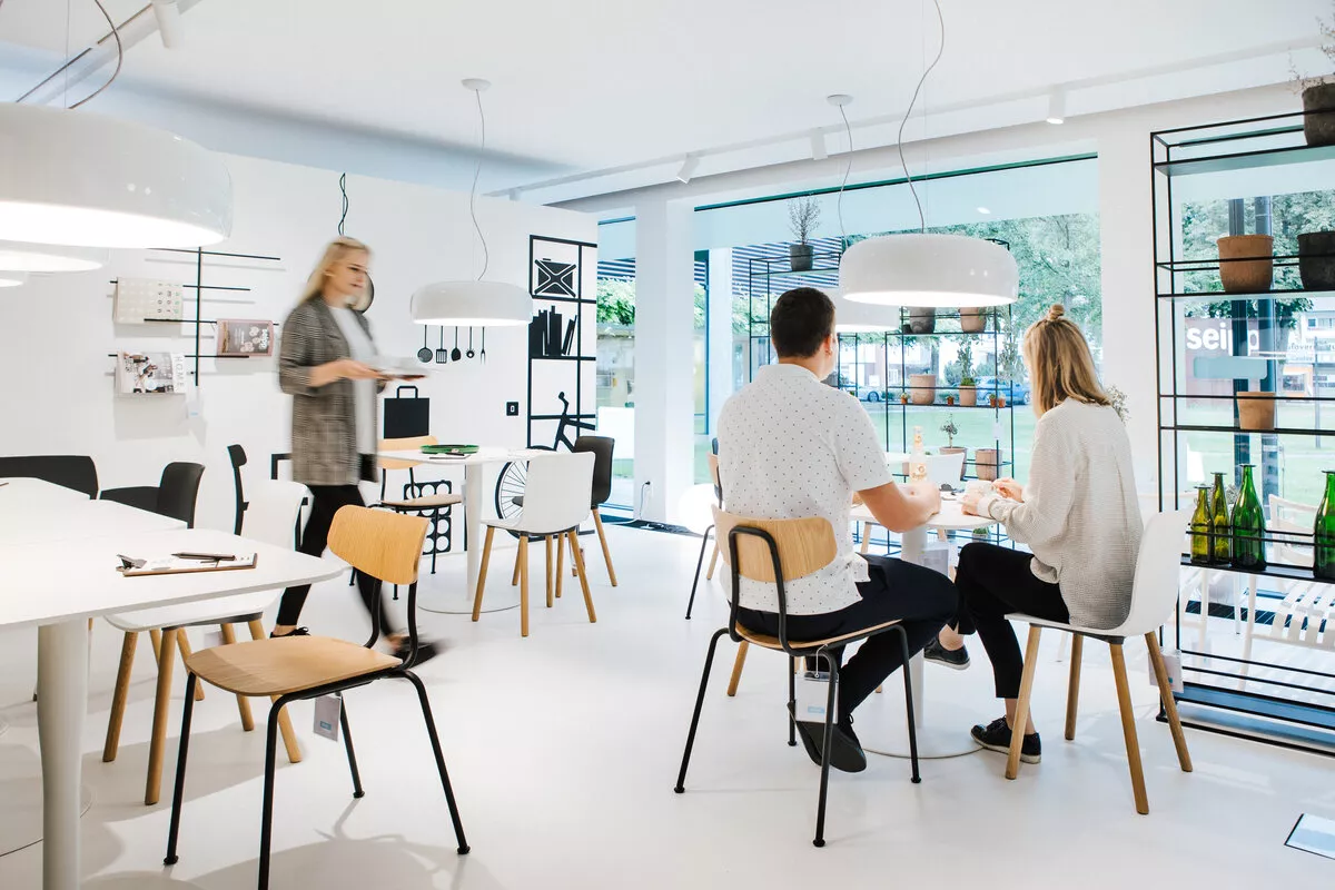 Innenaufnahme Café Super Normal mit Gästen, weißen Tischen, Holzstühlen und Fensterfront im Hintergrund. 