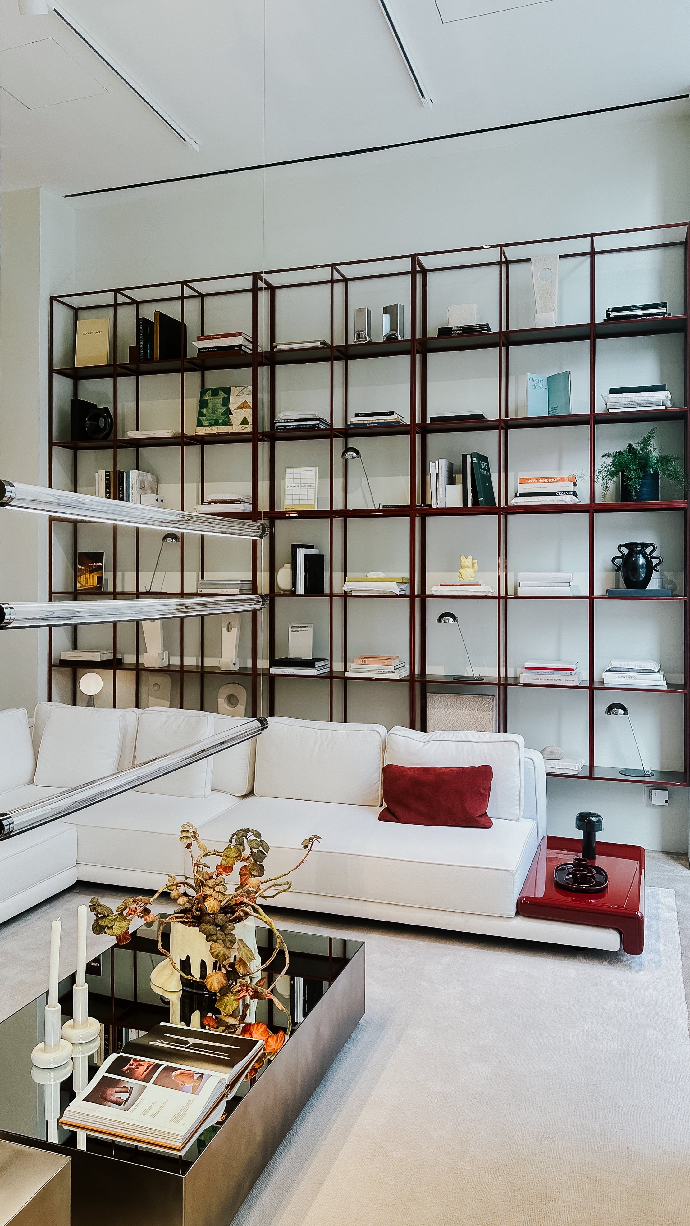Wohnszene mit wandfüllendem Regal aus feinen Metallprofilen, weißen Polstermöbeln und punktuellen Farbakzenten. Die Lichtführung und klaren Linien erzeugen eine ruhige, strukturierte Raumwirkung.