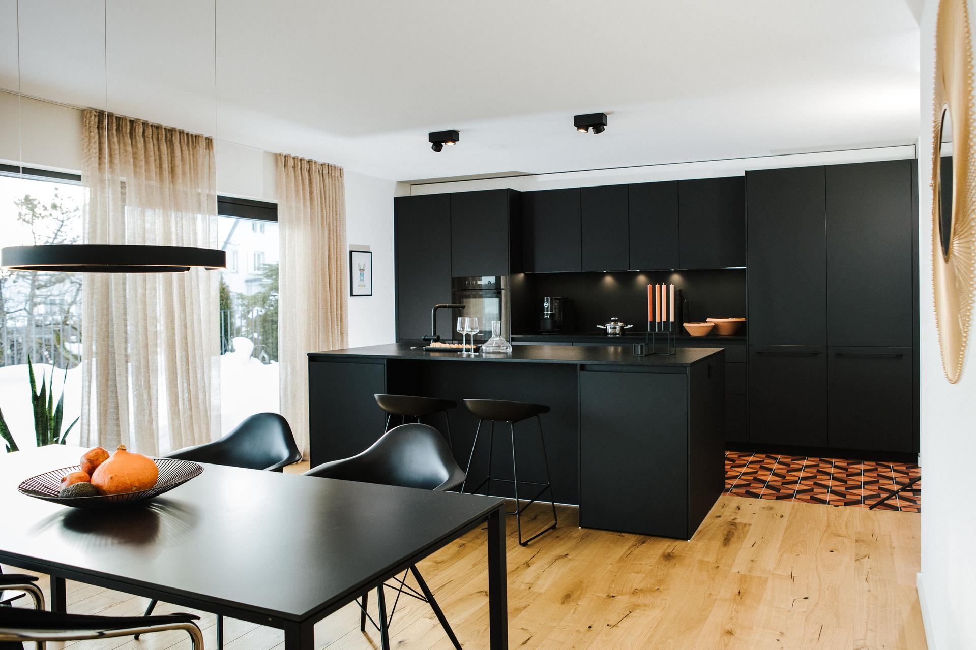 Küche in mattem Schwarz mit Kochinsel, Esstisch und Eames Chairs auf hellem Holzfußboden.  