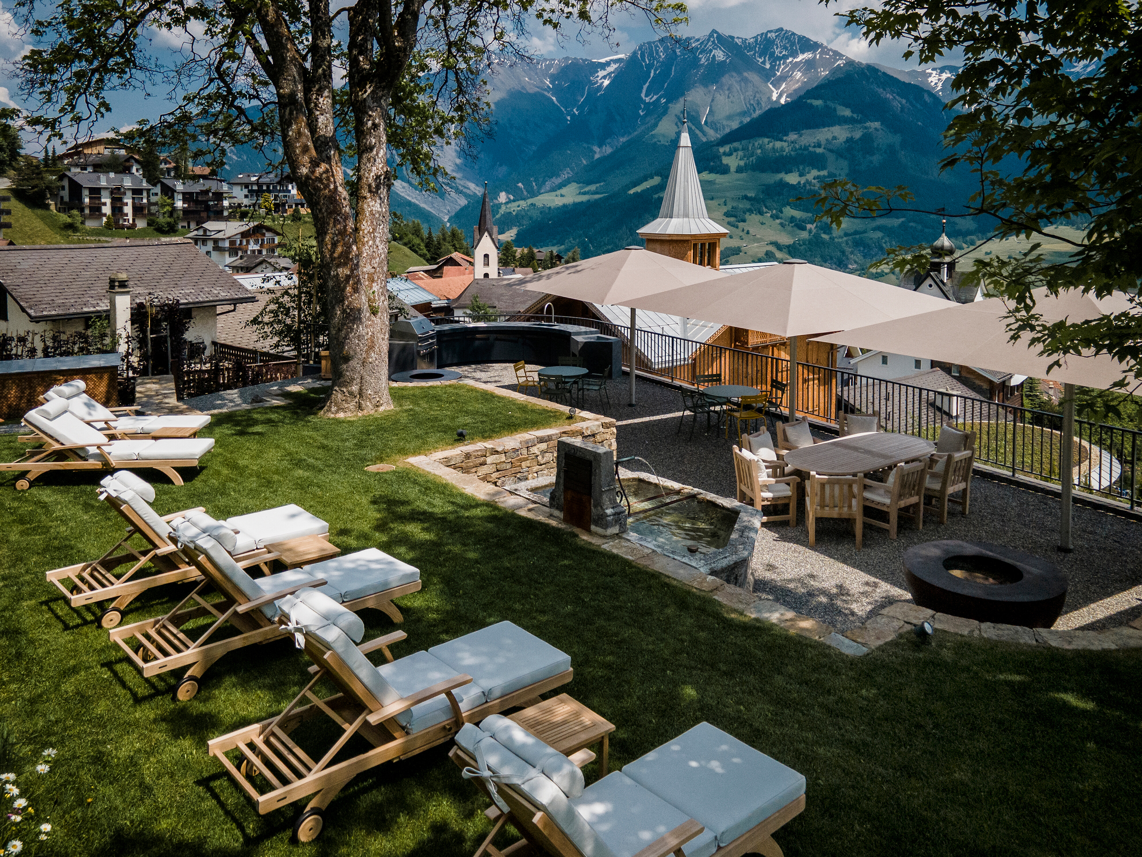 Liegen und Essbereich mit Sonnenschirmen auf einer Terrasse in Hanglage mit Bergblick und Dorfkulisse