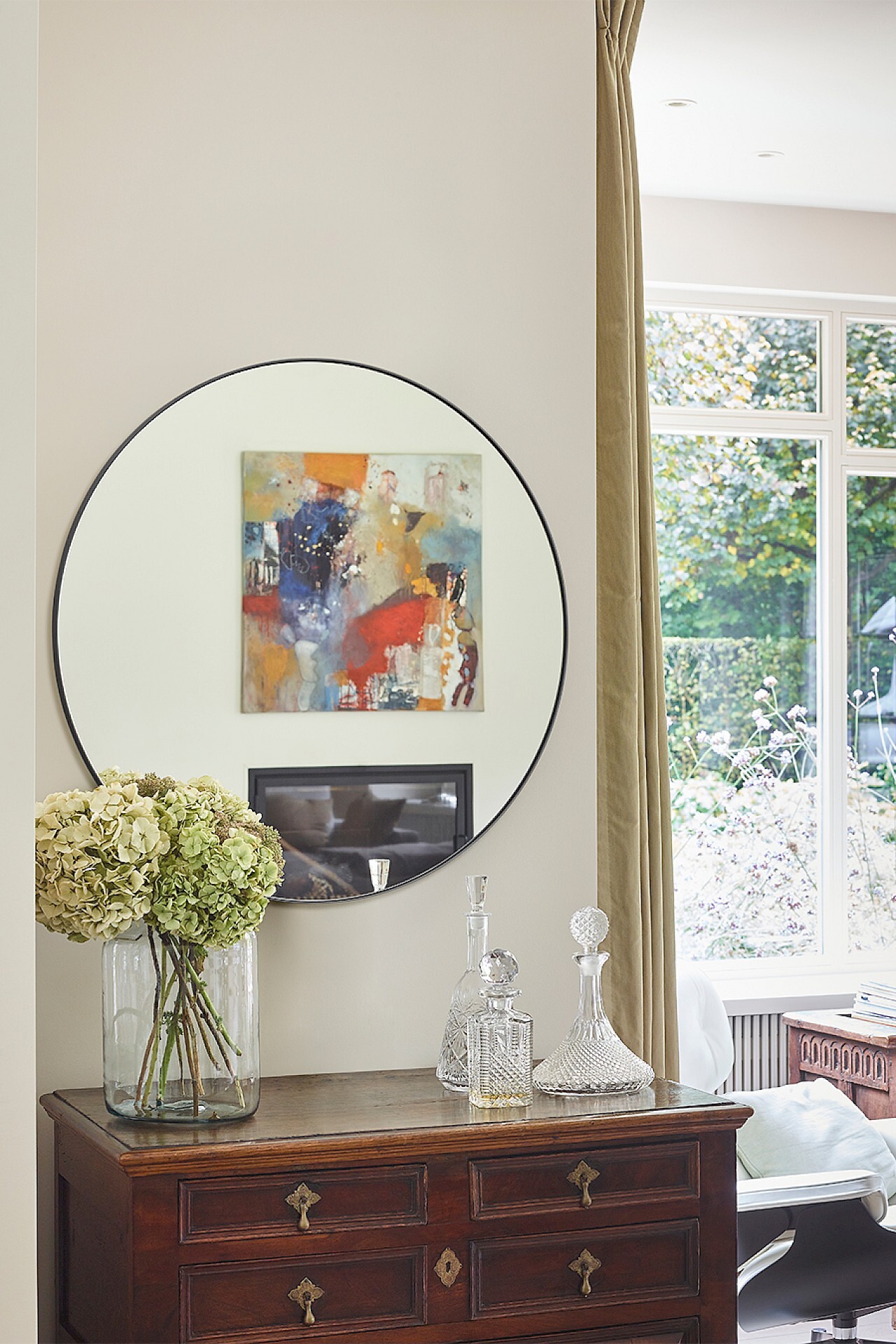 Runder Wandspiegel mit schmalem Rahmen über antiker Kommode mit Glasobjekten und Blumen