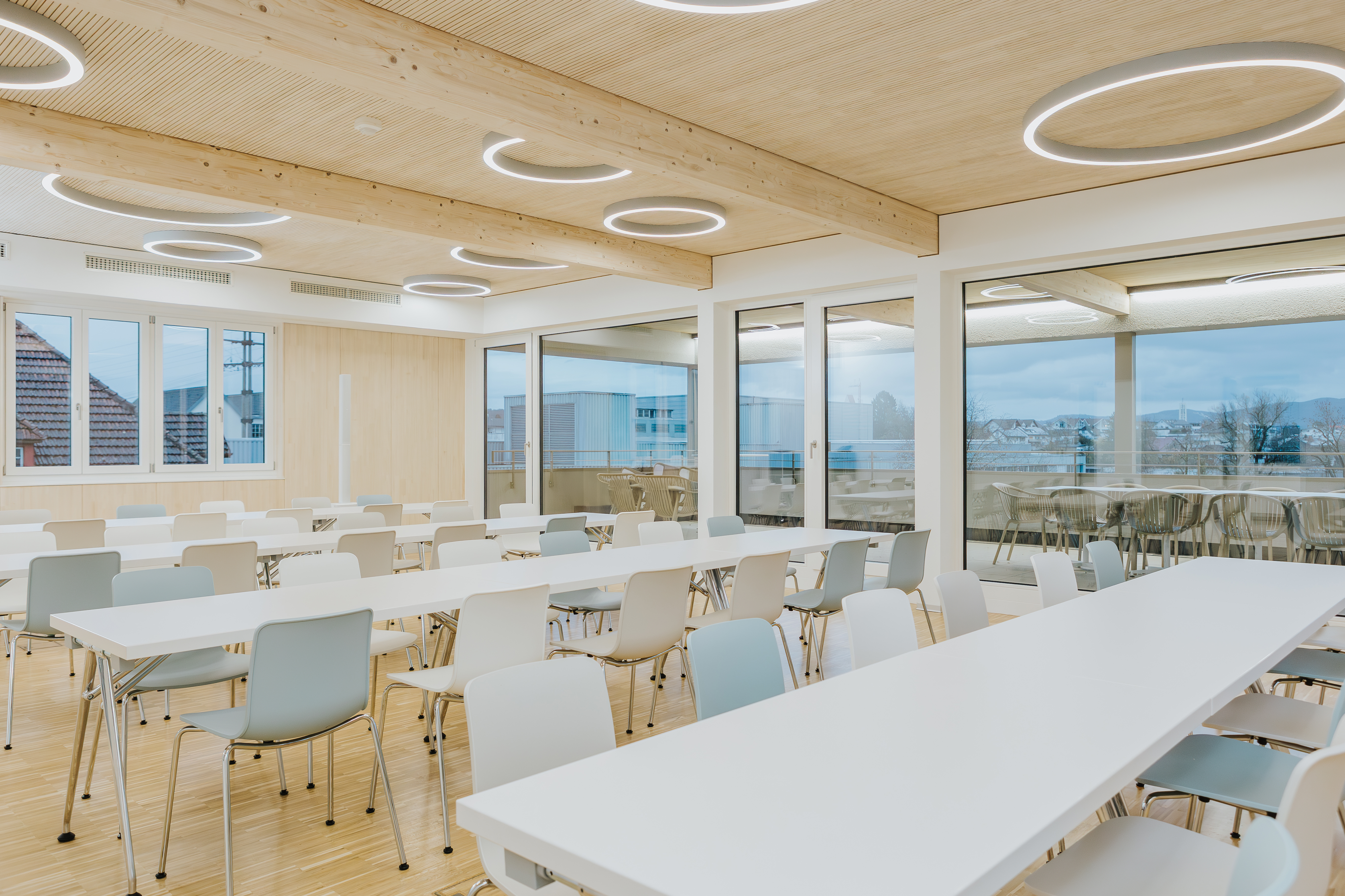 Großer, lichtdurchfluteter Aufenthaltsraum mit schlichten weißen Tischen und Stühlen in Pastelltönen.