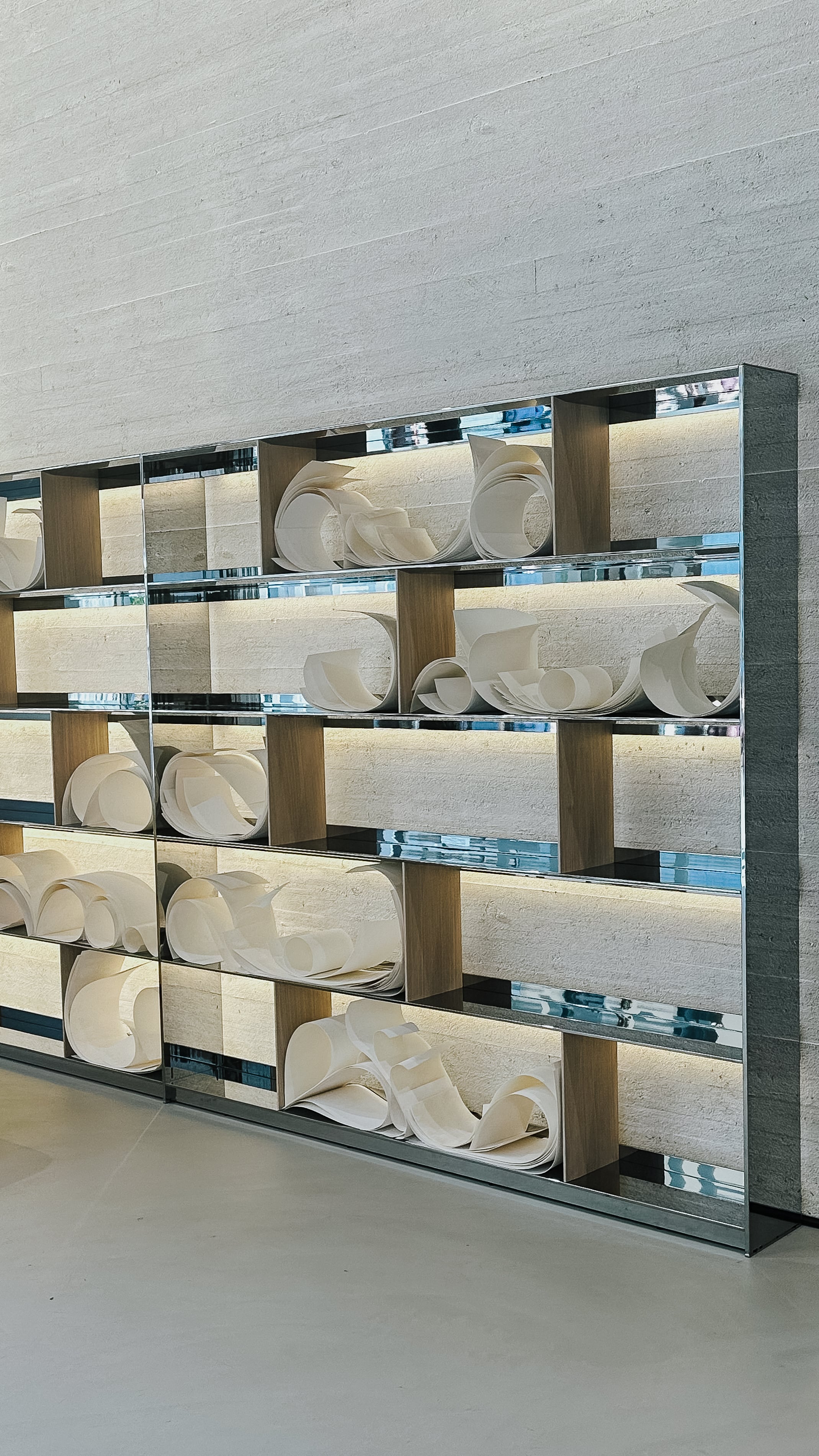 Skulpturales Regalsystem mit Chromstahl-Elementen, hinterleuchtet und bestückt mit abstrakten, papierartigen Objekten. Die glänzenden Oberflächen spiegeln Licht und Umgebung, betonen klare Linien und Materialästhetik.