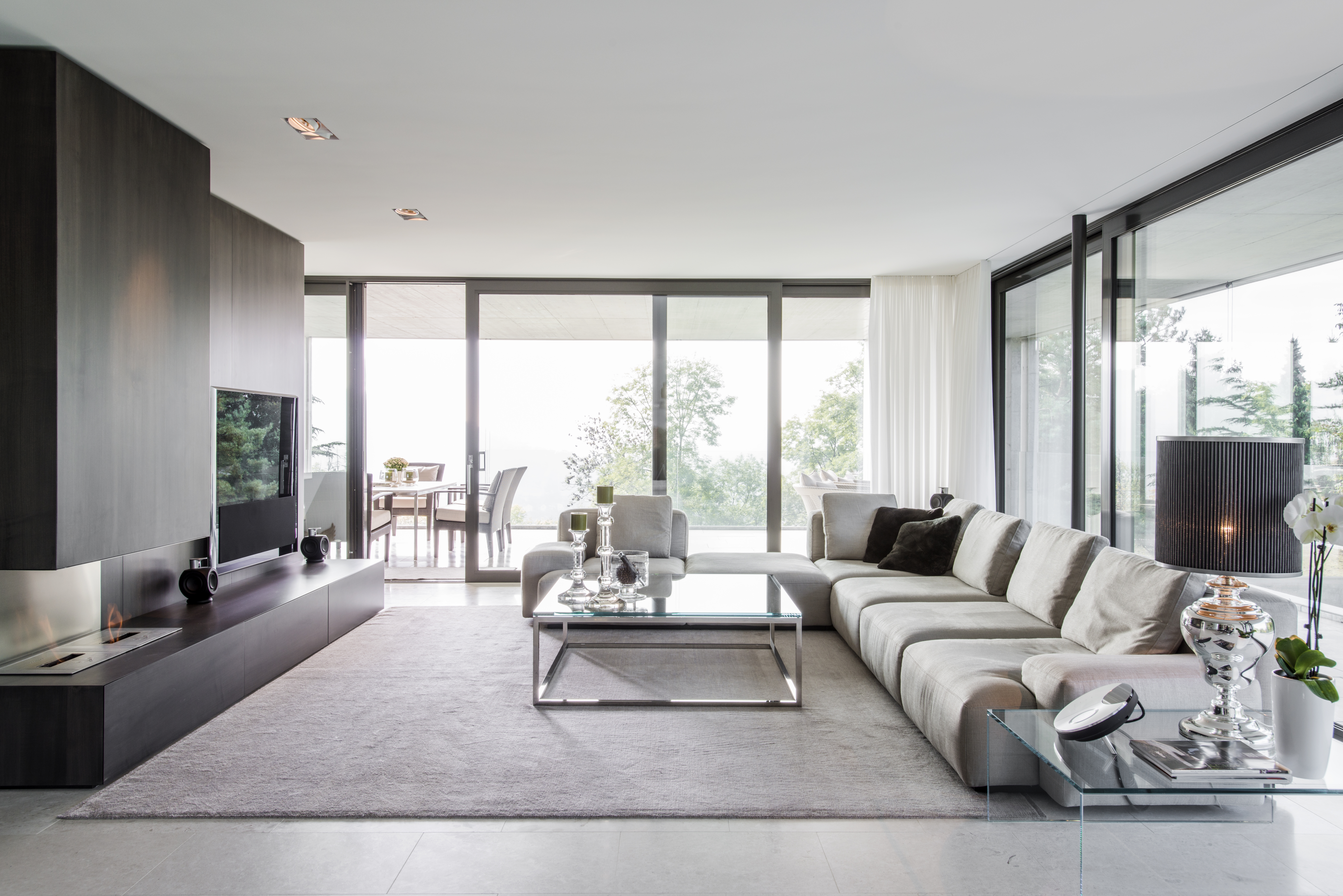 Wohnbereich mit bodentiefen Fenstern, grauem Ecksofa, Kaminwand in Dunkelholz und verchromten Details.