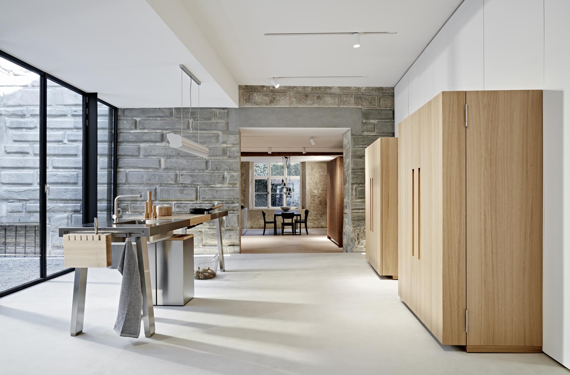 Bulthaup Küche mit freistehender Edelstahlinsel und Werkschränken in heller Holzoptik vor Steinwand 