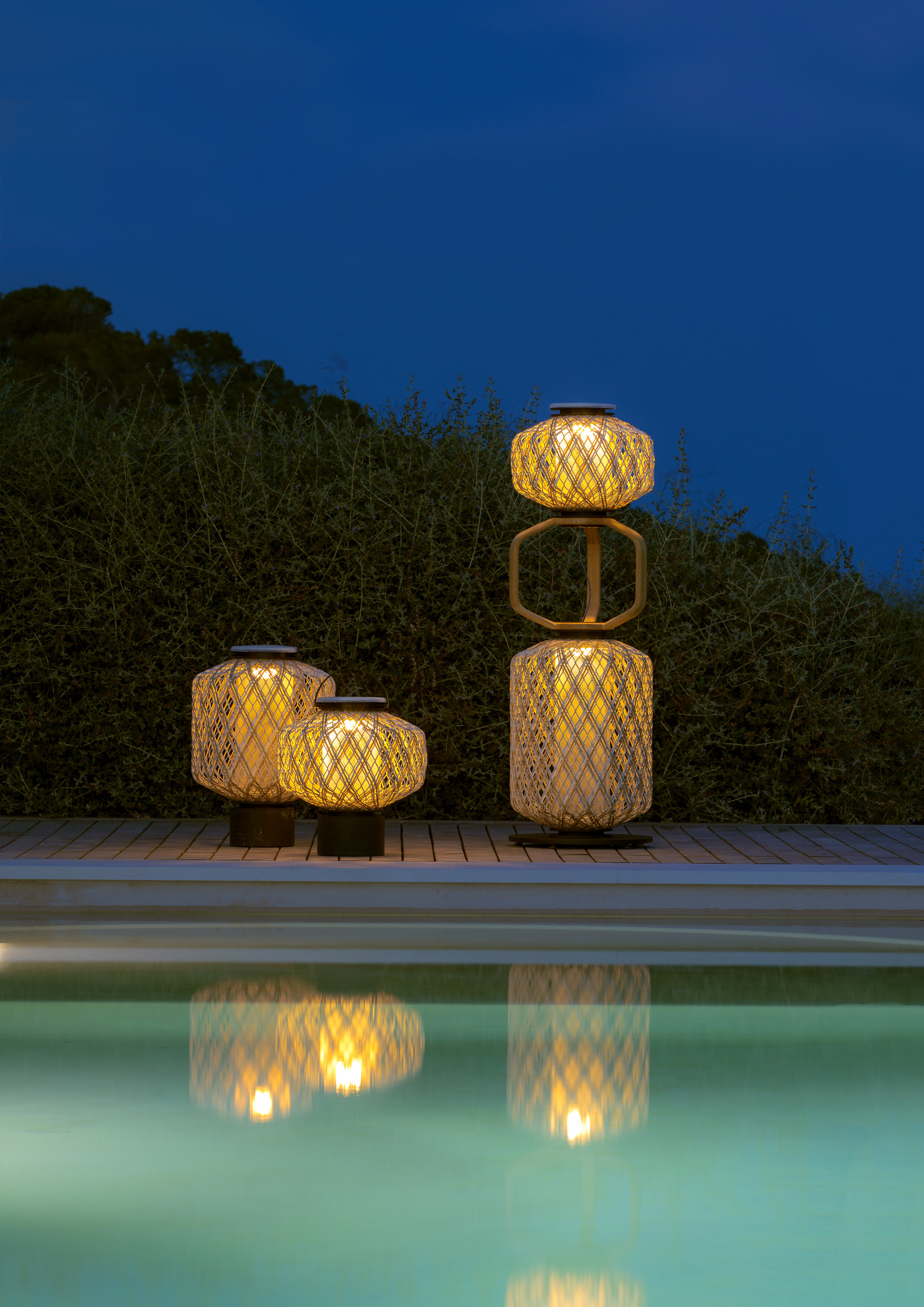 Verschiedene Outdoor-Leuchten aus Flechtmaterial auf Holzdeck mit Bepflanzung im Hintergrund