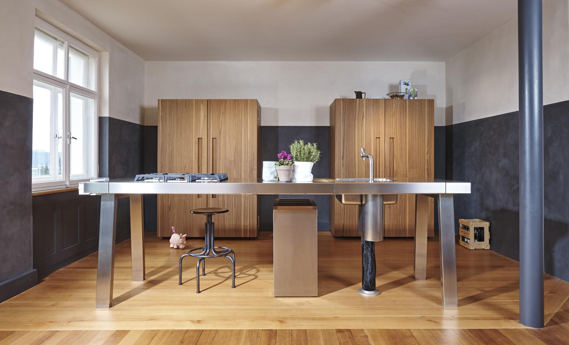 Offene Küche mit bulthaup b2 Edelstahlinsel, Hockern und Werkschränken aus markantem Holz. 