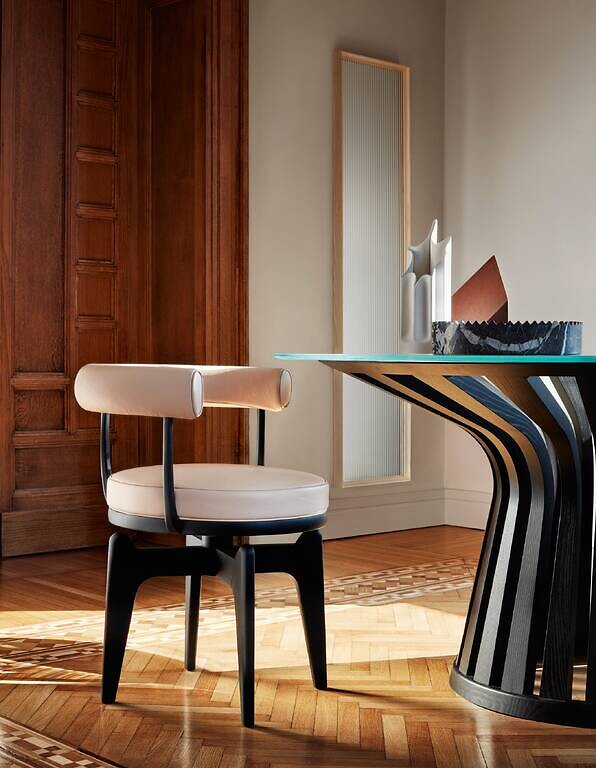 Stuhl mit gepolsterter Rückenrolle an Glastisch mit skulpturalem Gestell auf Parkettboden  