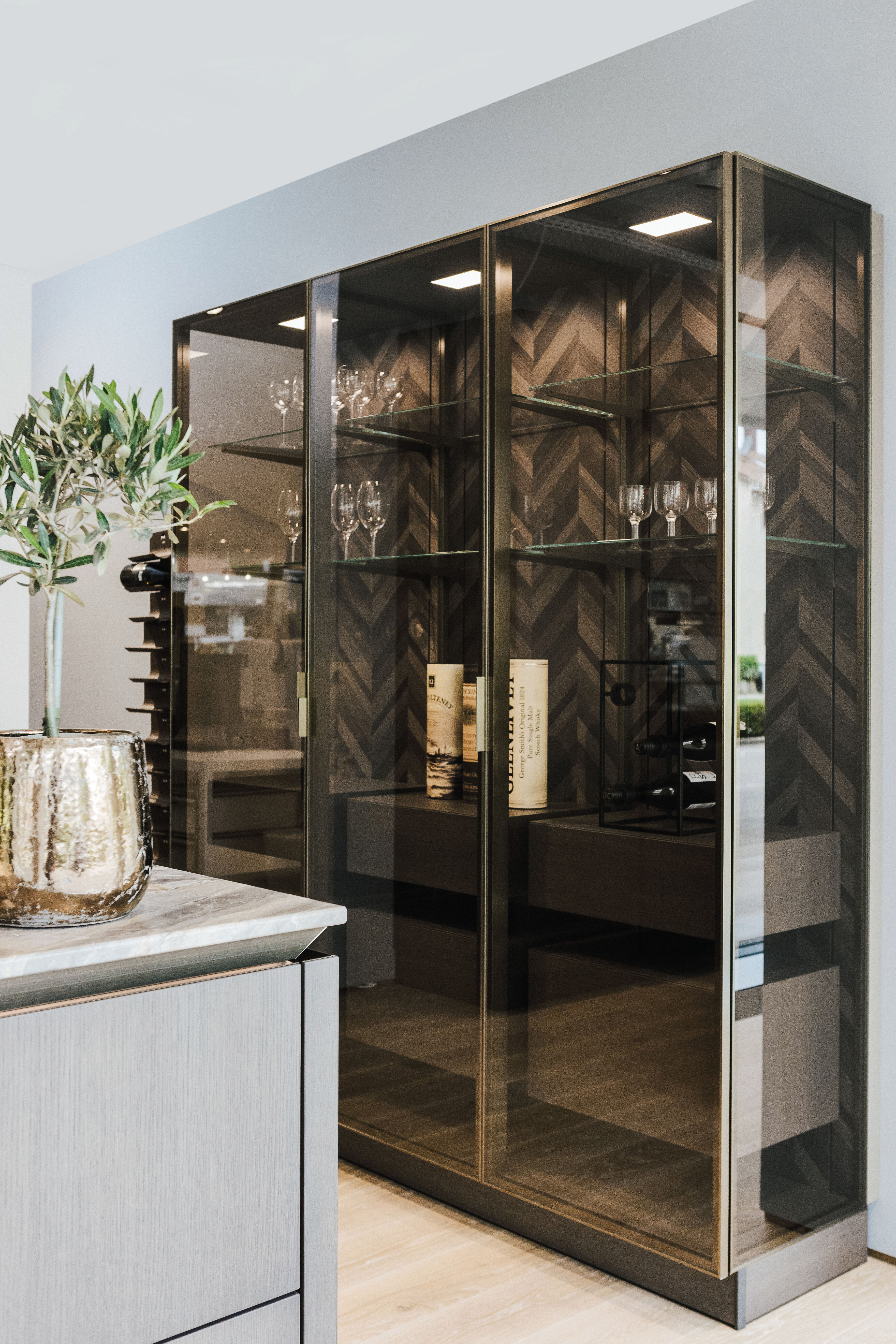 Vitrinenschrank mit Glasfront und dunkler Holzrückwand in Fischgrätoptik, innen mit LED-Beleuchtung