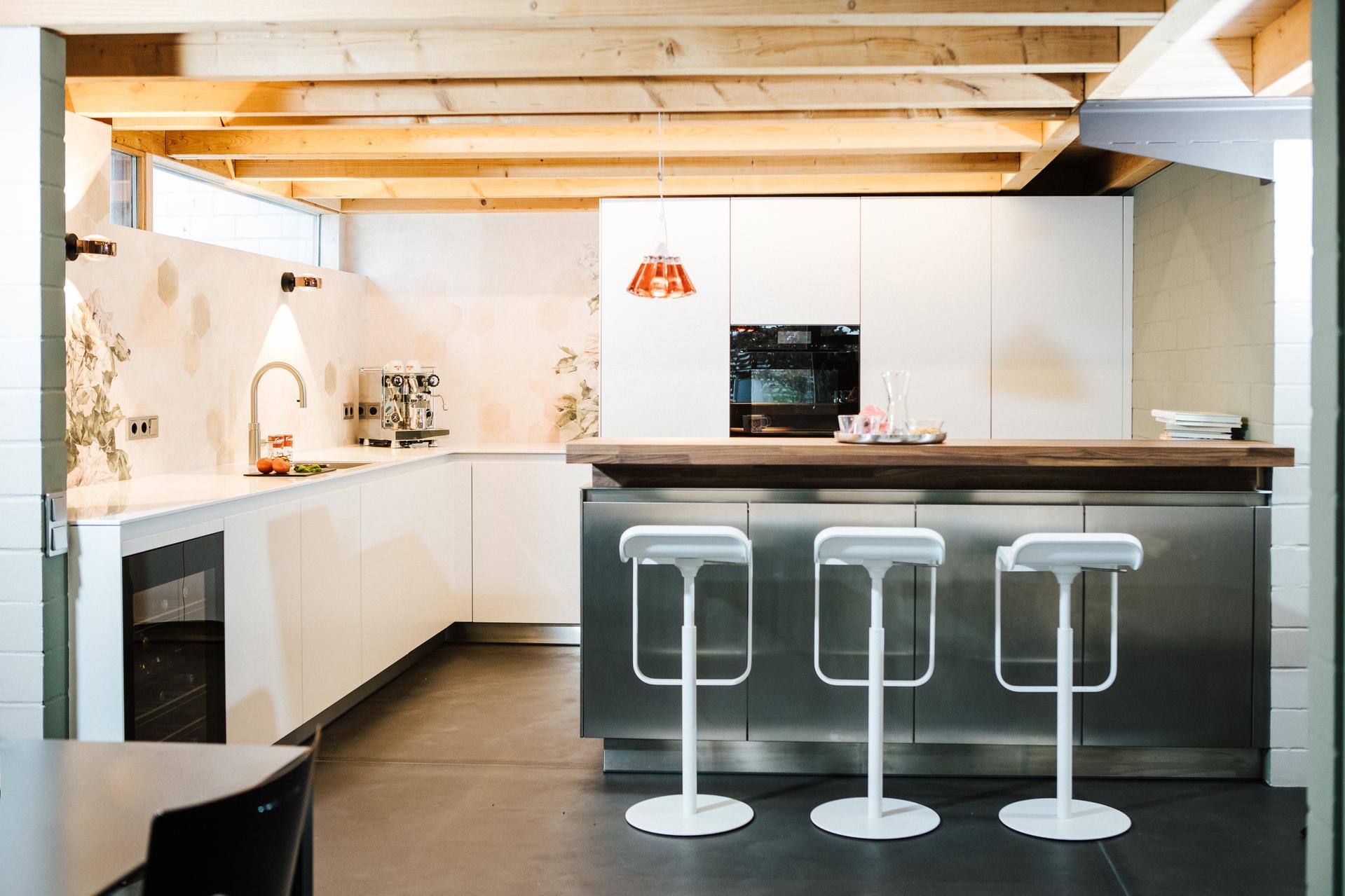 Moderne Küche mit weißer L-Form-Einrichtung, Kochinsel mit Holzarbeitsplatte, drei weißen Barhockern und freiliegenden Holzbalken an der Decke.