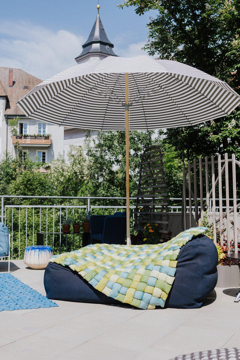 Großer Sitzsack mit grün-blauer Flechtdecke unter einem gestreiften Sonnenschirm auf der Dachterrasse von Seipp Wohnen in Waldshut