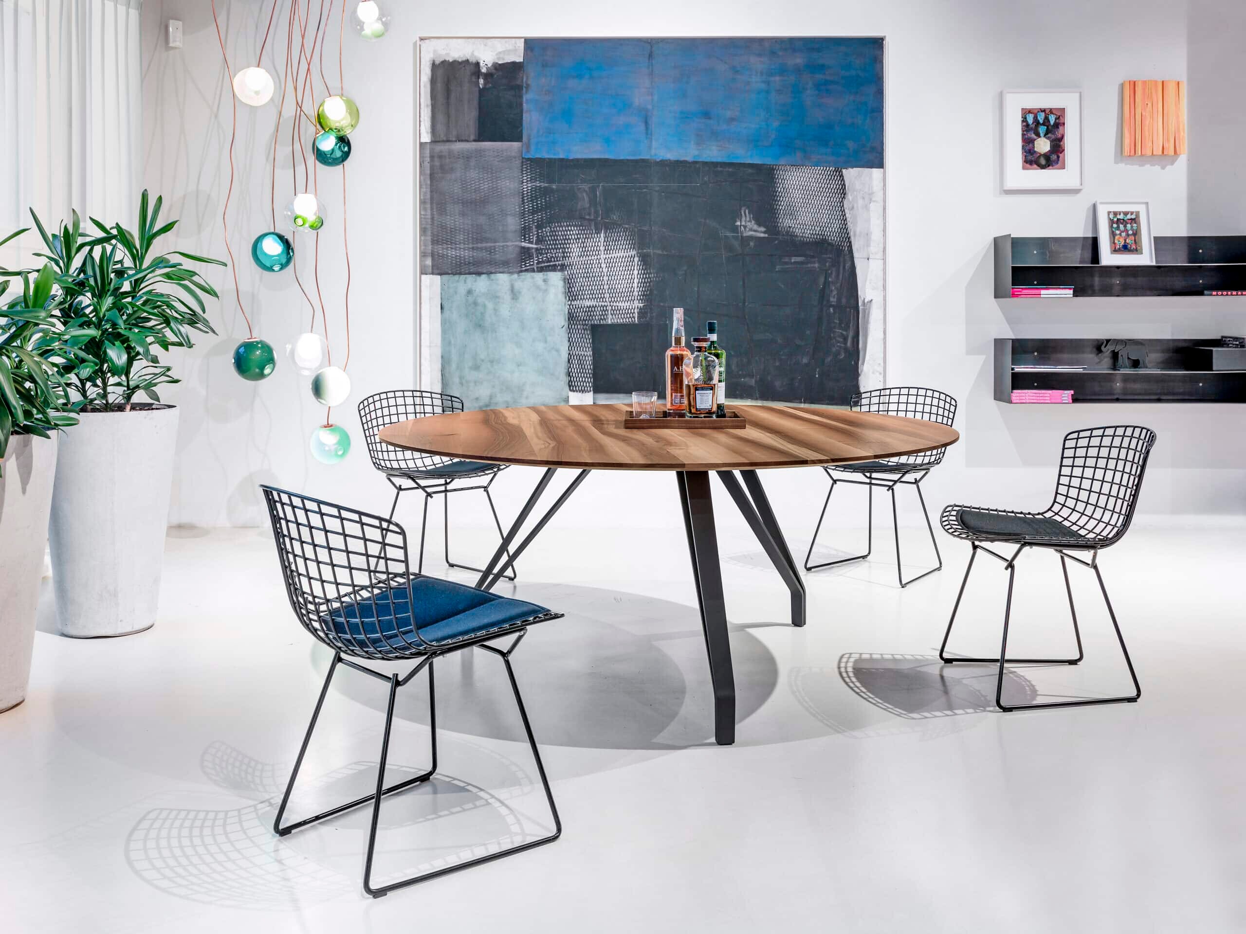 Vier filigrane Drahtgitterstühle und runder Holztisch vor farbig strukturierter Wand