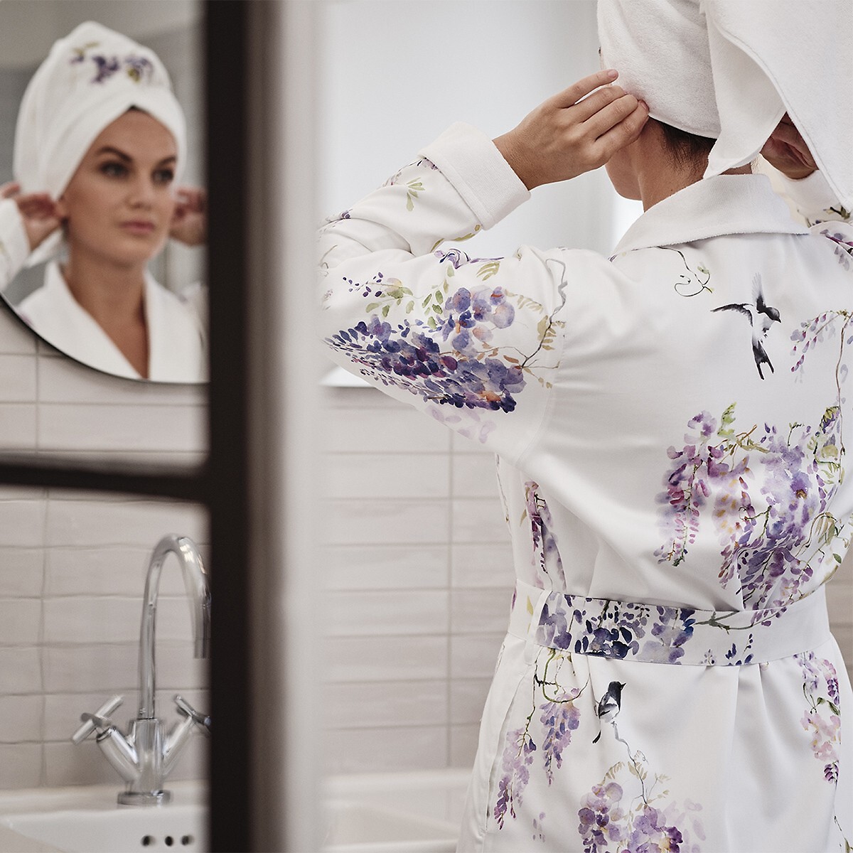 Frau im geblümten Morgenmantel steht mit Turban vor einem Spiegel im Bad