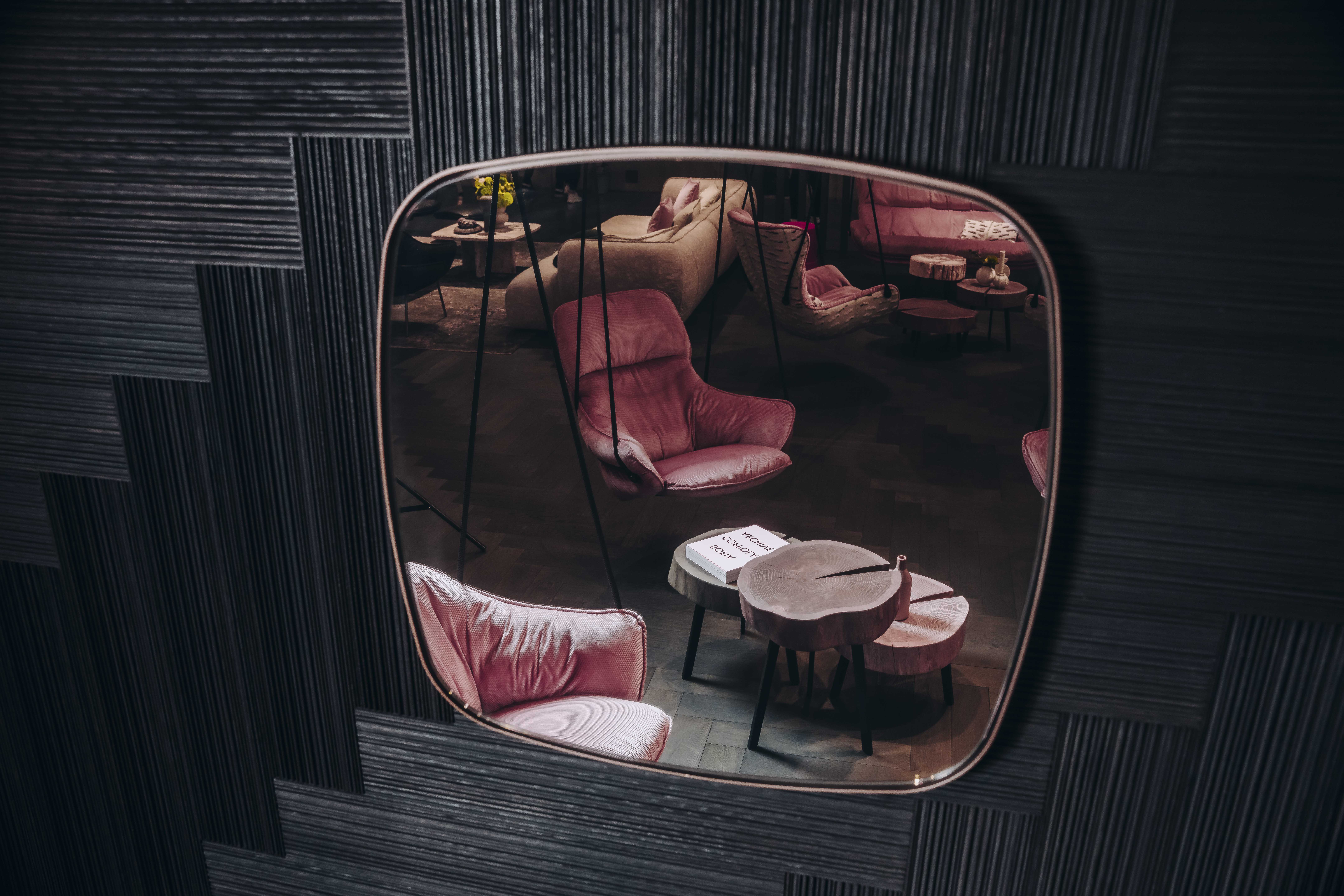 Spiegelaufnahme einer Messeinszenierung von Janua mit skulpturalen Holztischen, rosafarbenen Polstersesseln und markanter Wandverkleidung aus Massivholzpaneelen – sichtbar detailverliebt inszeniert.