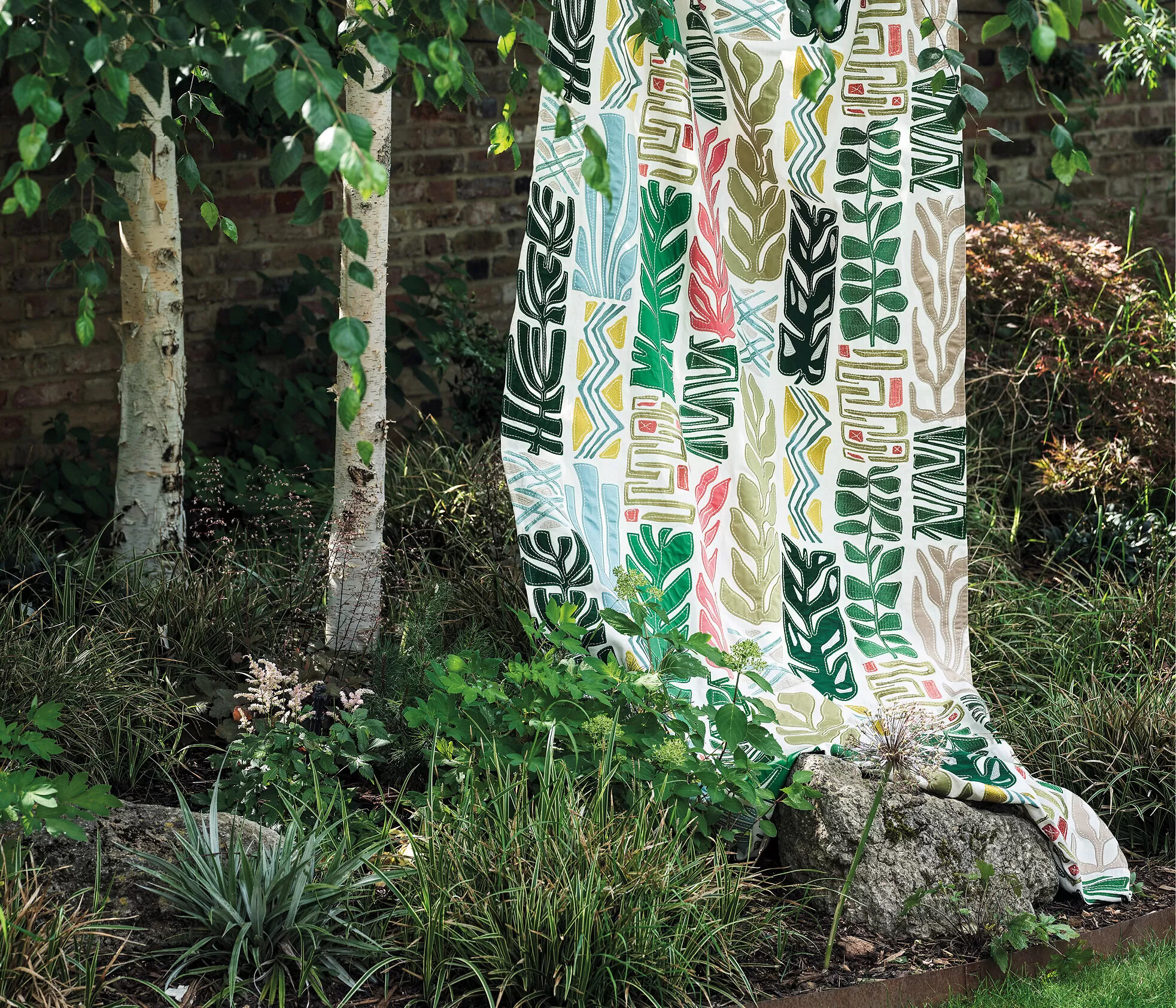 Gemusterter Vorhangstoff mit Blattmotiven hängt im Garten neben Birken und Sträuchern  