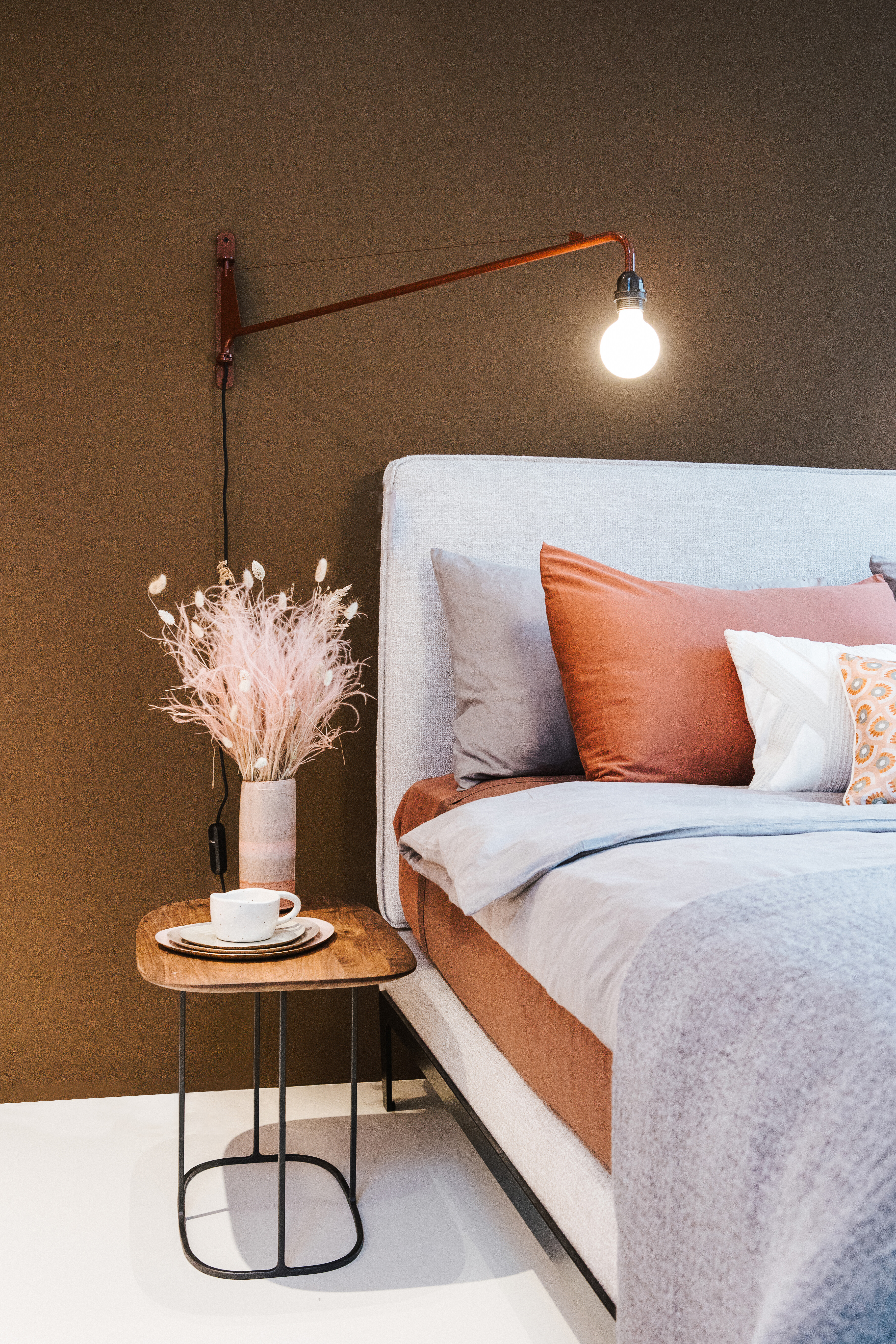 Detailaufnahme eines Bettes mit Wandlampe, Holz-Beistelltisch und dekorativem Trockenstrauß