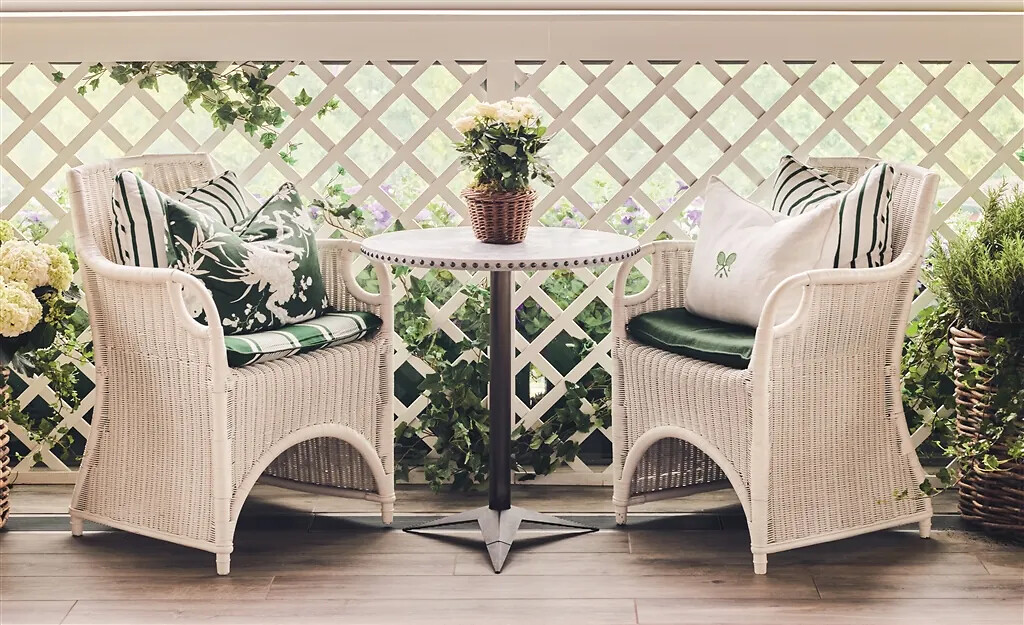 Zwei weiße Flechtstühle mit grün gemusterten Kissen und runder Tisch auf bepflanztem Balkon  