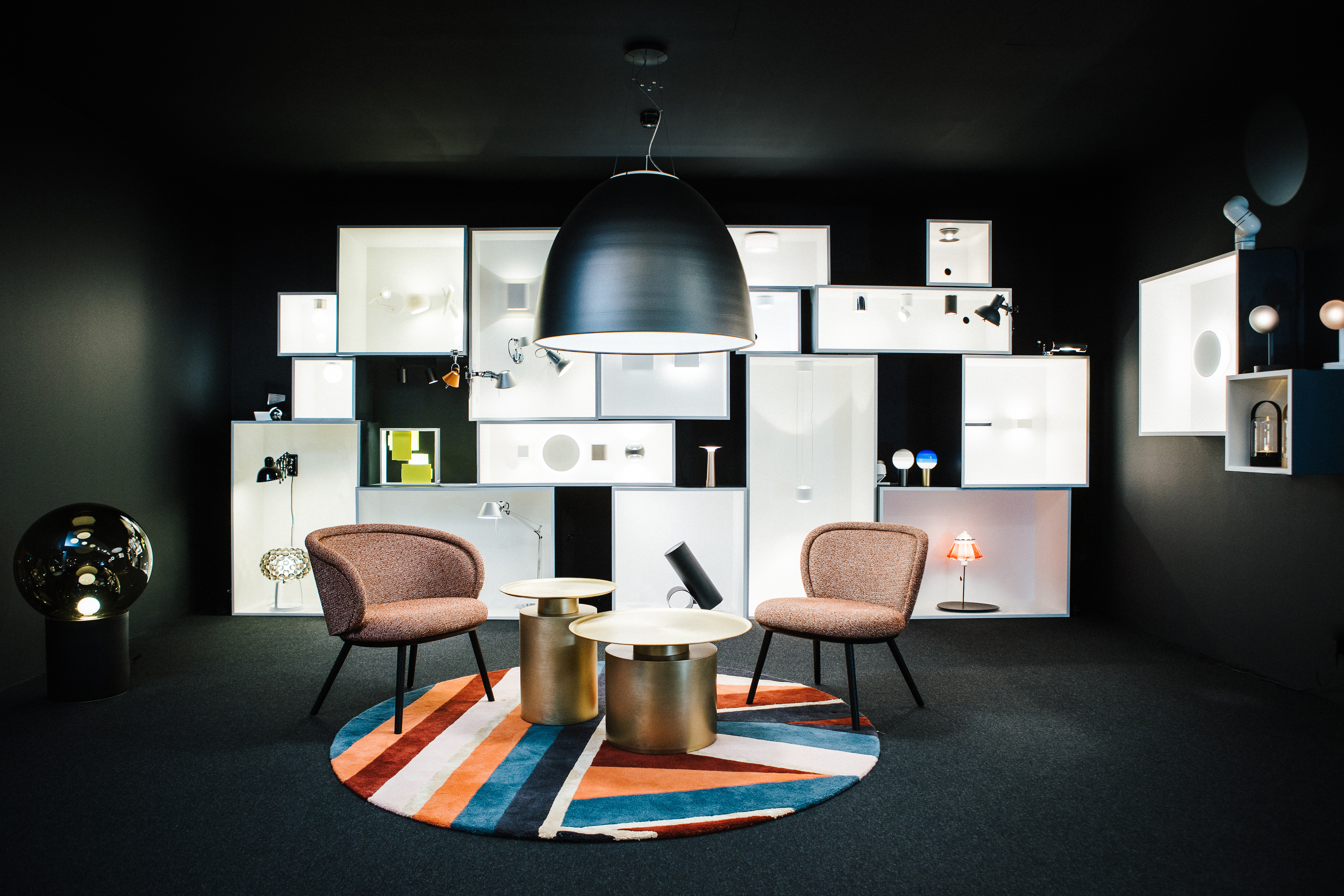 Ausstellungsraum mit schwarzen Wänden,  beleuchteten Nischen, zwei Sesseln und Beistelltischen auf farbigem Teppich