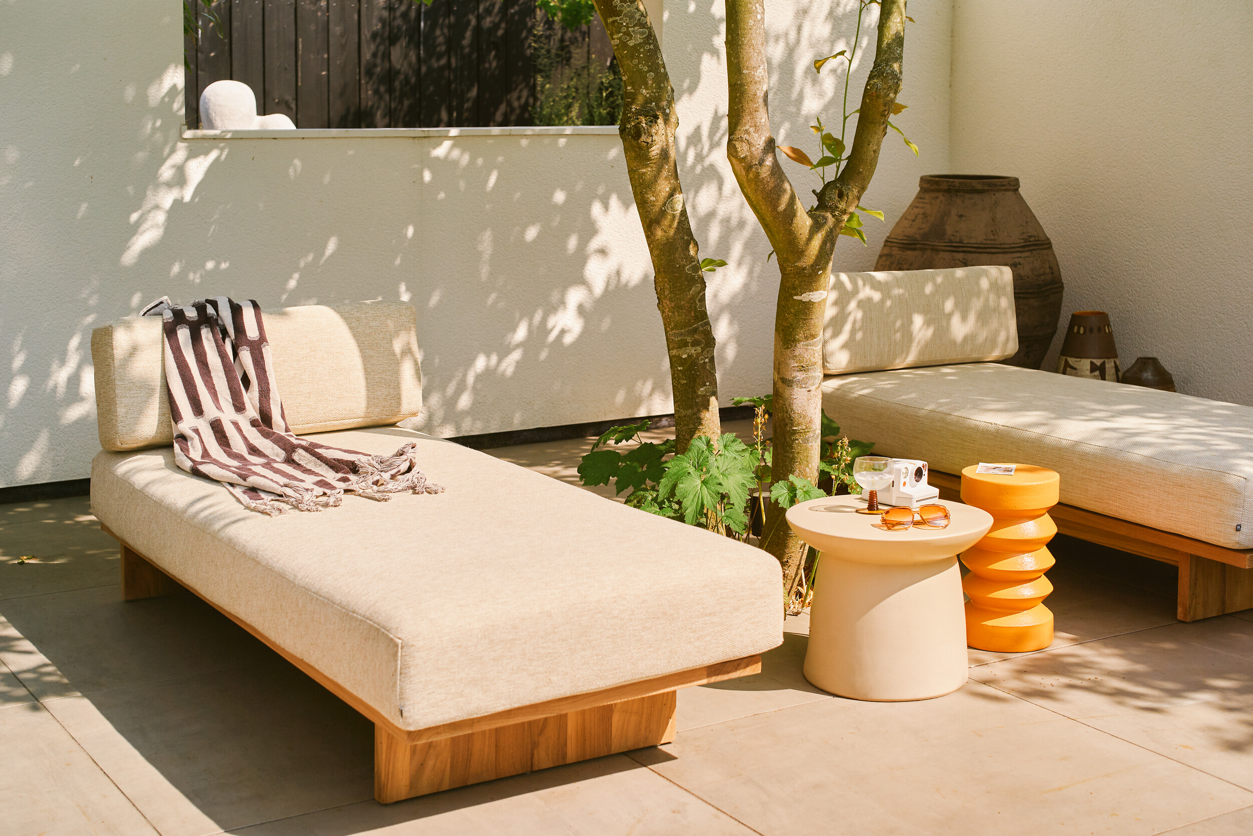 Zwei Daybeds mit hellem Bezug und orangefarbenem Beistelltisch unter Baum auf Terrasse  