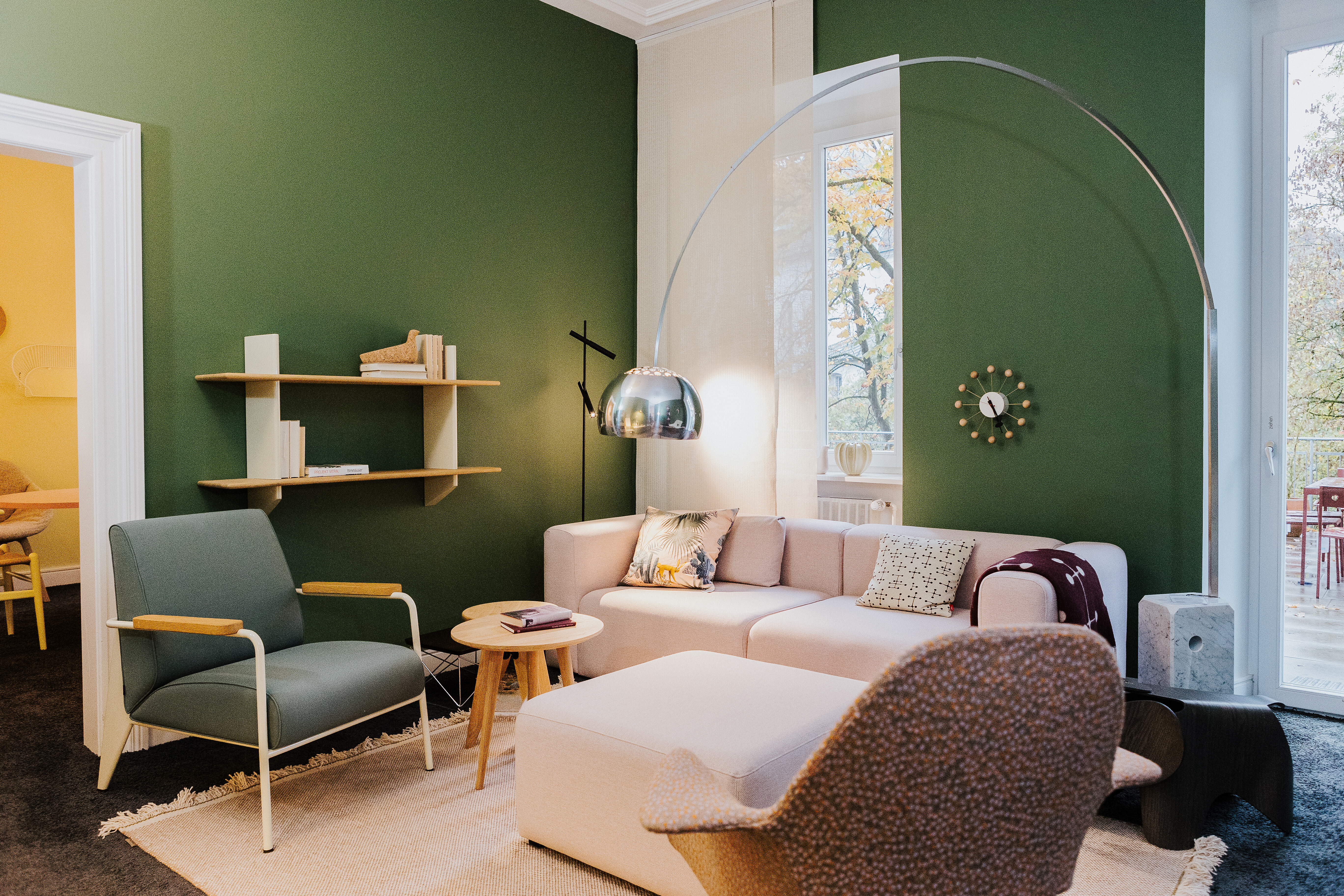 Wohnbereich mit grünem Wandanstrich, hellen Polstermöbeln, Bogenleuchte und Holzregal an der Wand