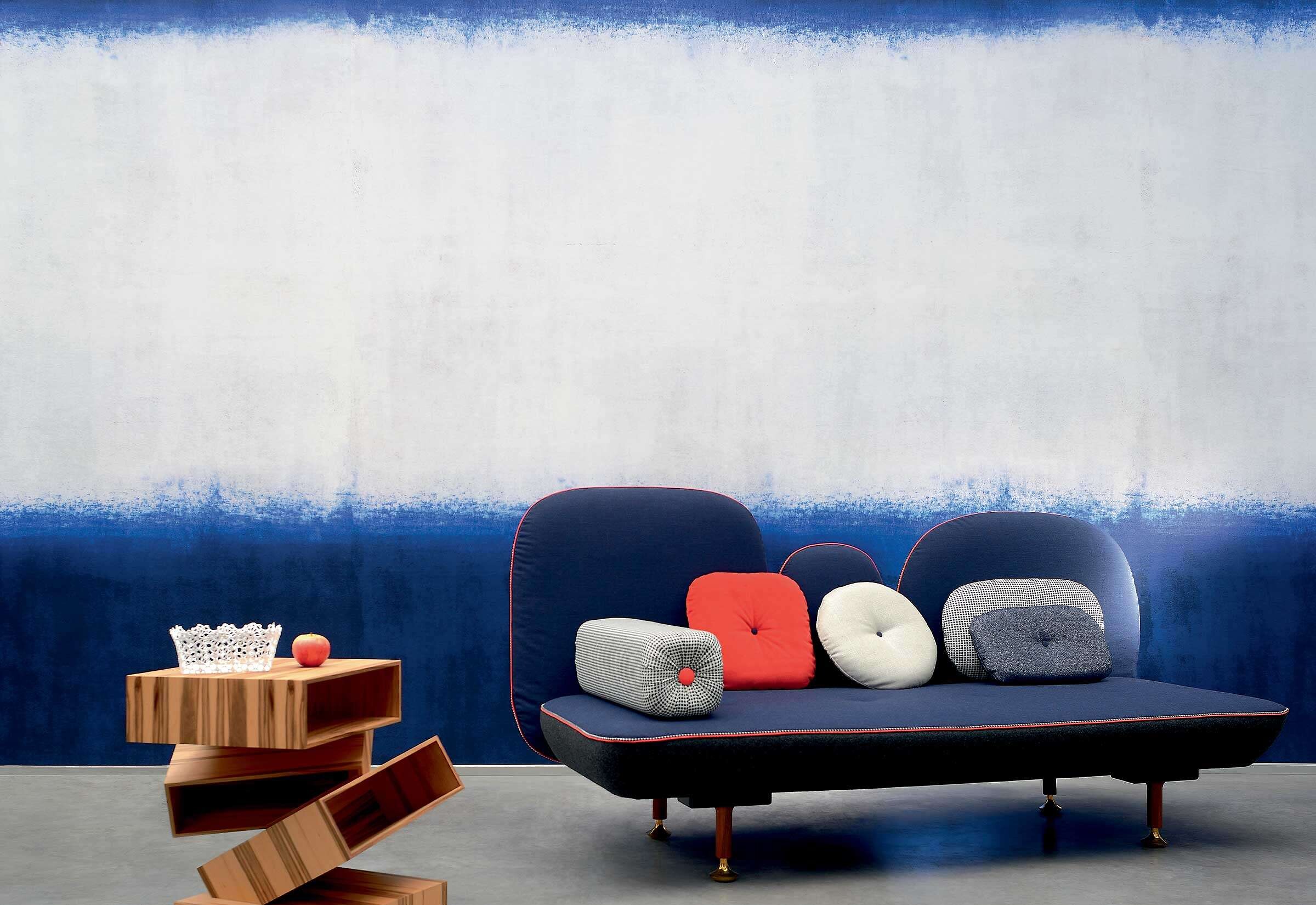 Blaues Sofa mit runden Kissen vor Wand mit Farbverlauf von Weiß zu Blau und skulpturalem Beistelltisch