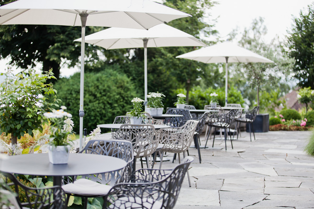 Terrasse mit filigranen Gartenstühlen, runden Tischen und weißen Sonnenschirmen in grüner Umgebung.