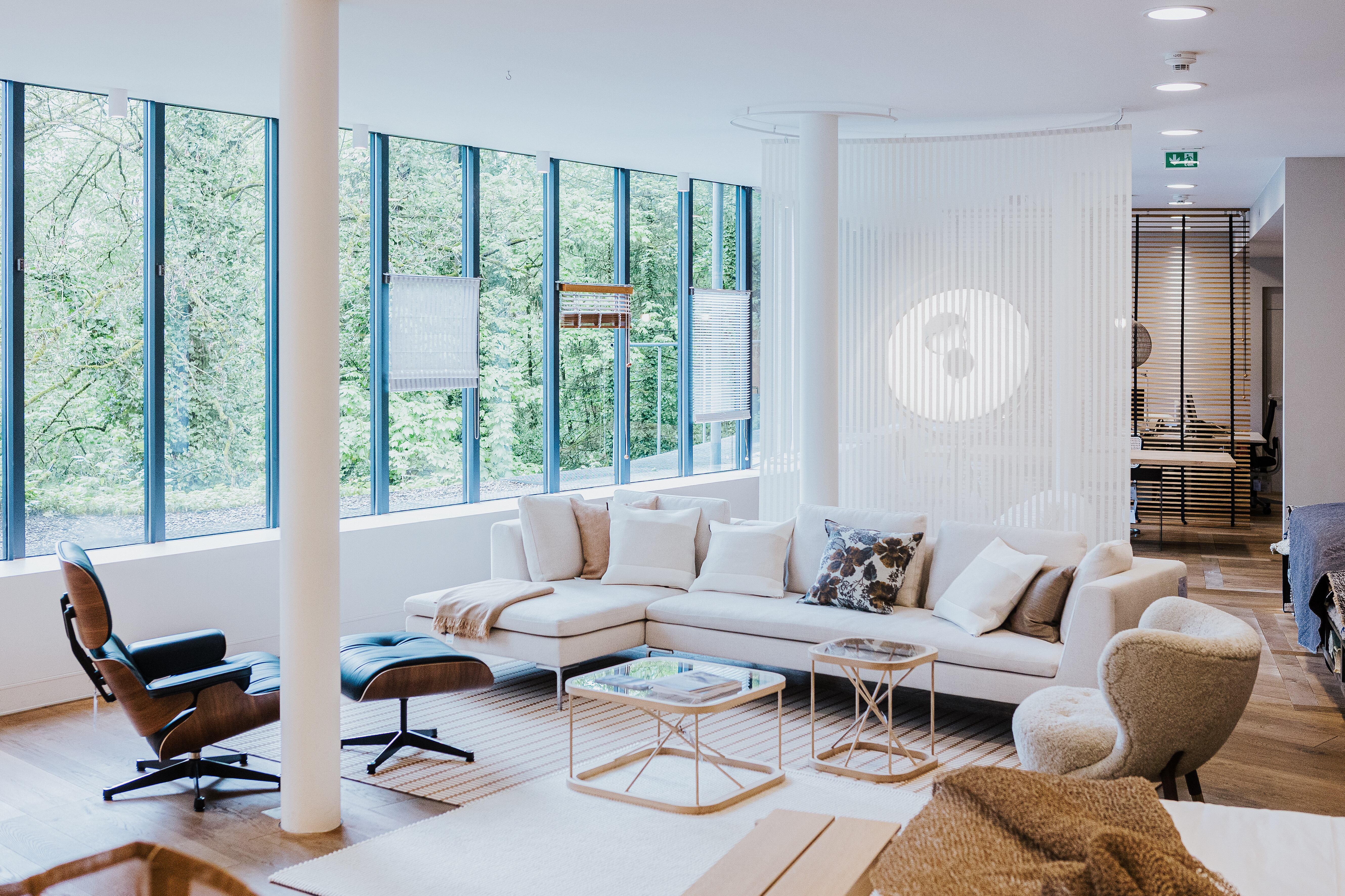 Wohnbereich mit heller Sofalandschaft, Eames Lounge Chair und raumhohen Fenstern mit Waldblick