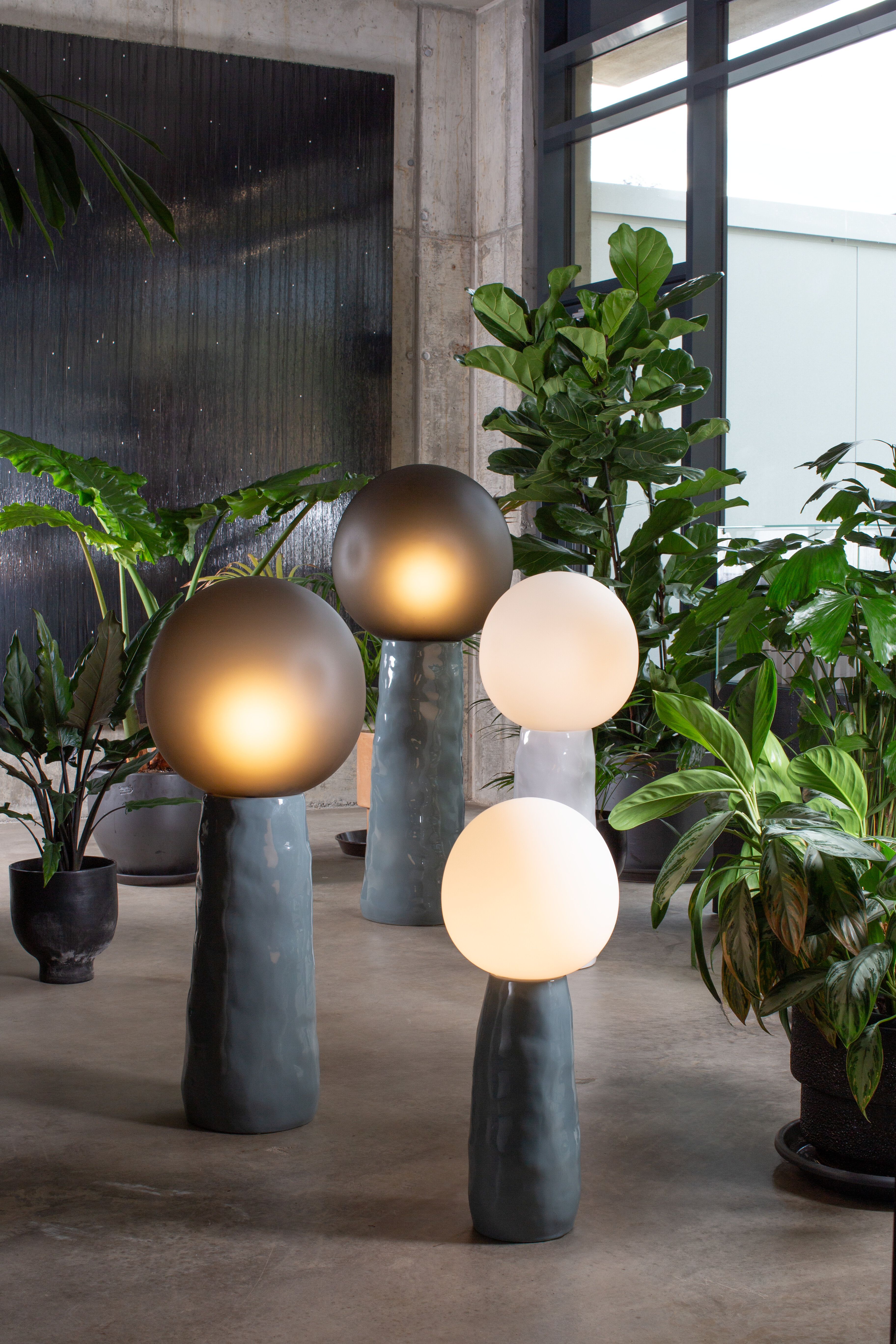 Drei Stehleuchten mit kugelförmigem Leuchtkörper auf strukturiertem Sockel zwischen Pflanzen  