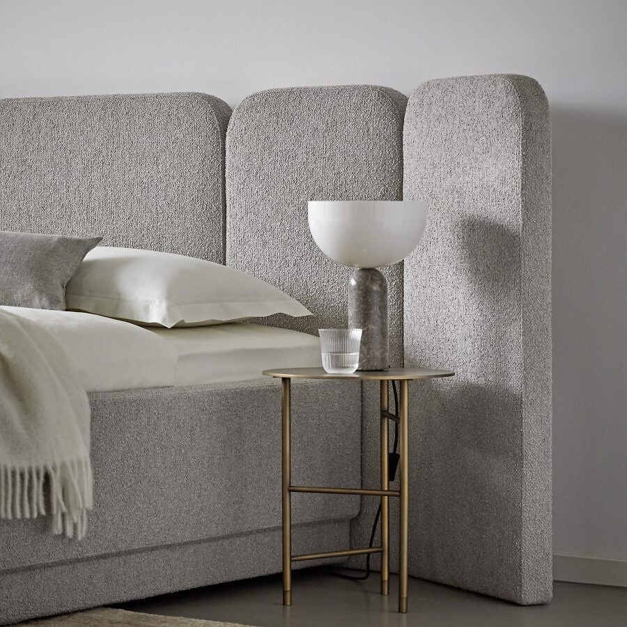 Bett mit grauem Polsterkopfteil, Plaid und Leuchte auf Beistelltisch