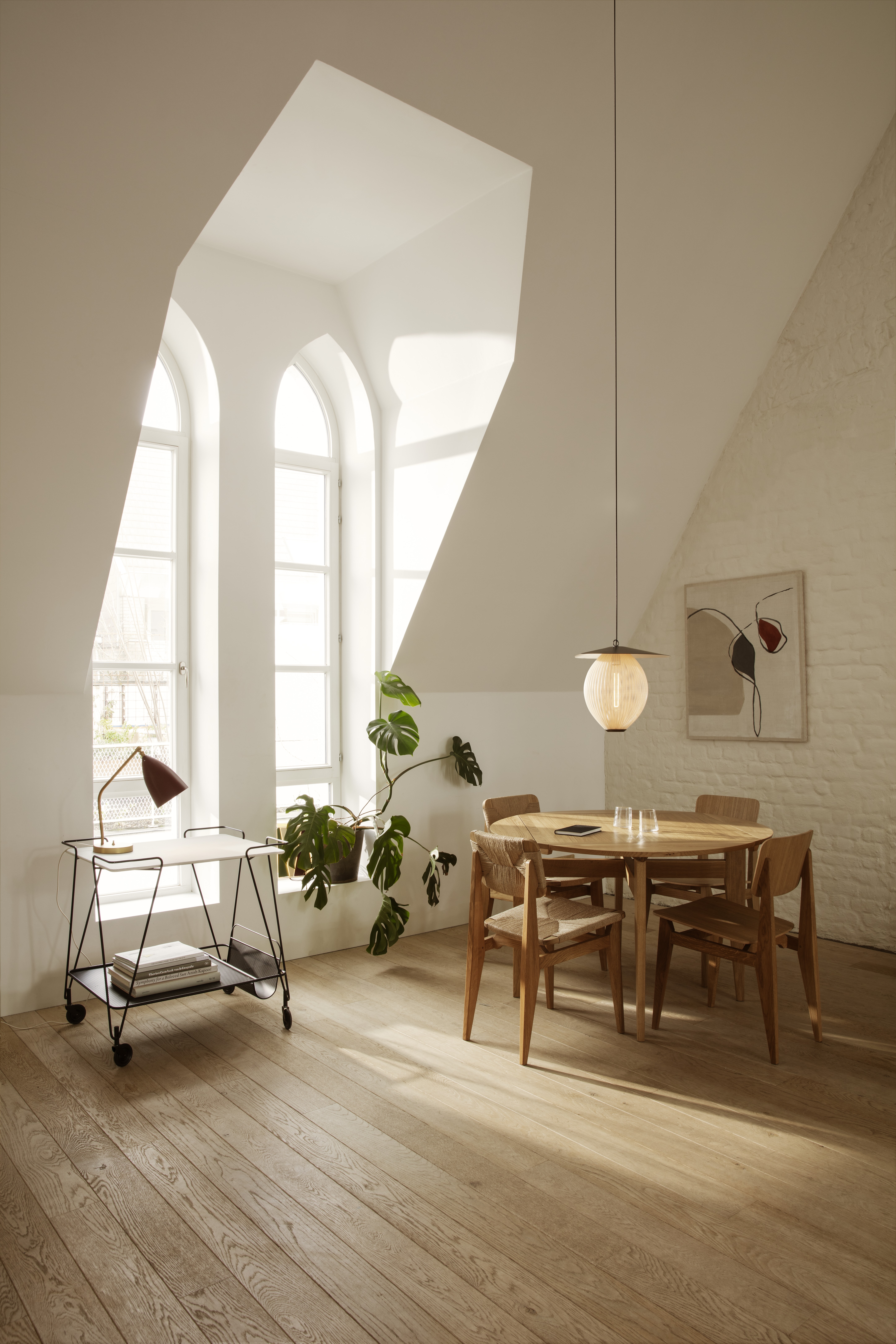 Runder Holztisch mit vier Stühlen, Pendelleuchte und Servierwagen vor hohen Fenstern mit Spitzbogen