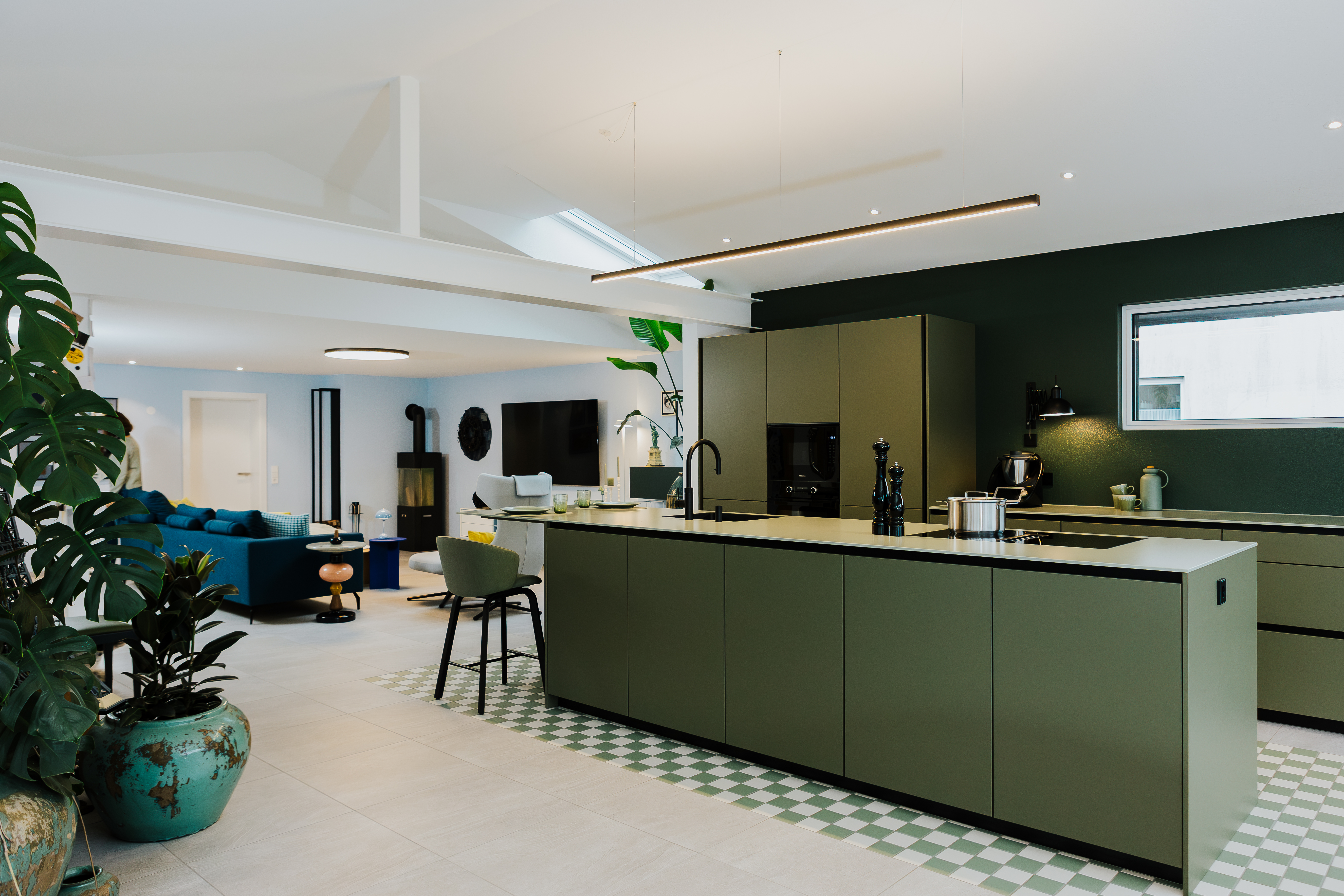 Offene Wohnküche mit olivgrüner Insel, grün akzentuierter Wand und markanten Pflanzen im Raum.
