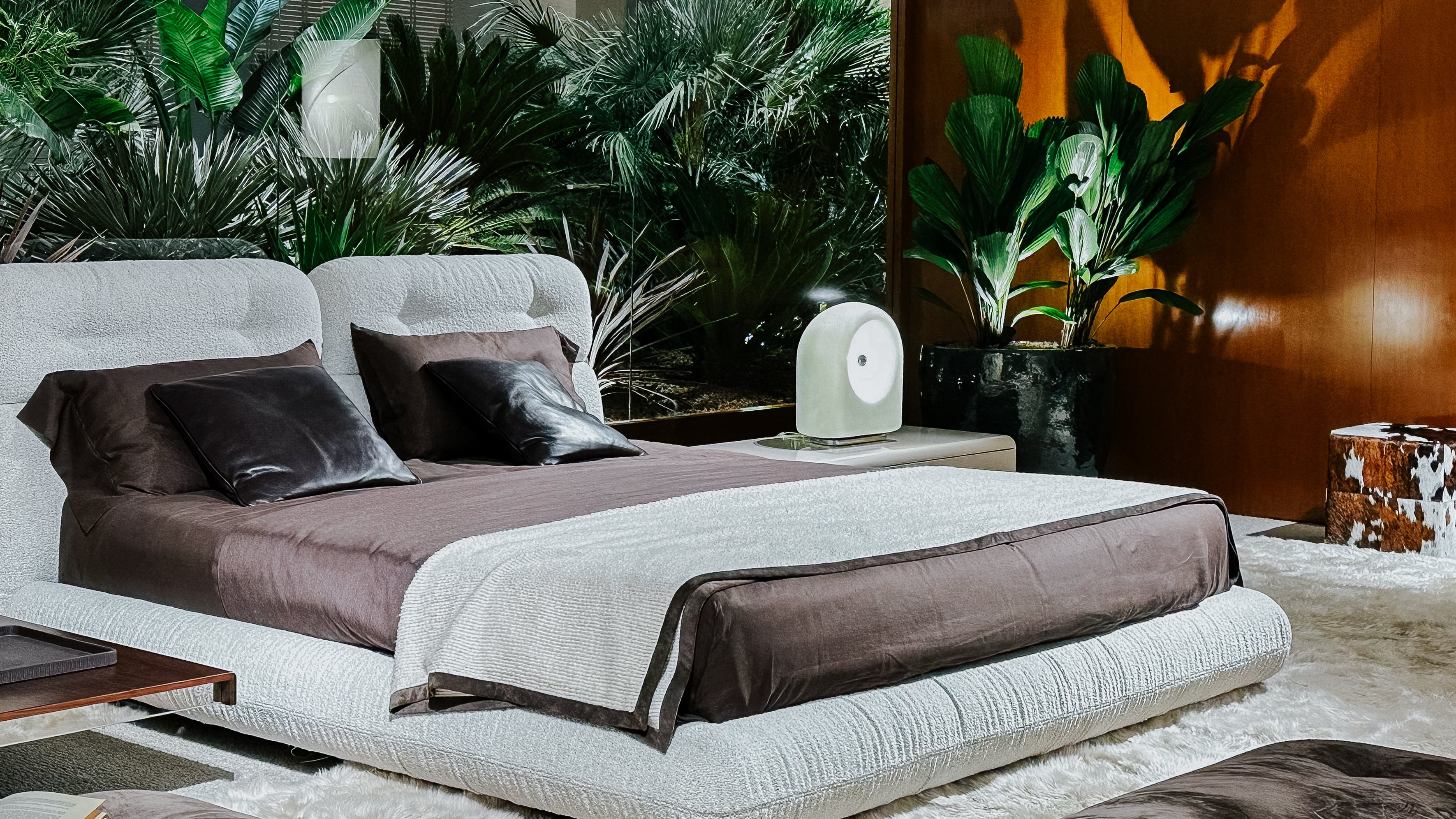 Designbett mit Bouclé-Bezug und brauner Bettwäsche in einer Messeszene auf der Il Salone del Mobile 2025. Umgeben von großblättrigen Pflanzen, Holzverkleidung und weichem Teppichboden.