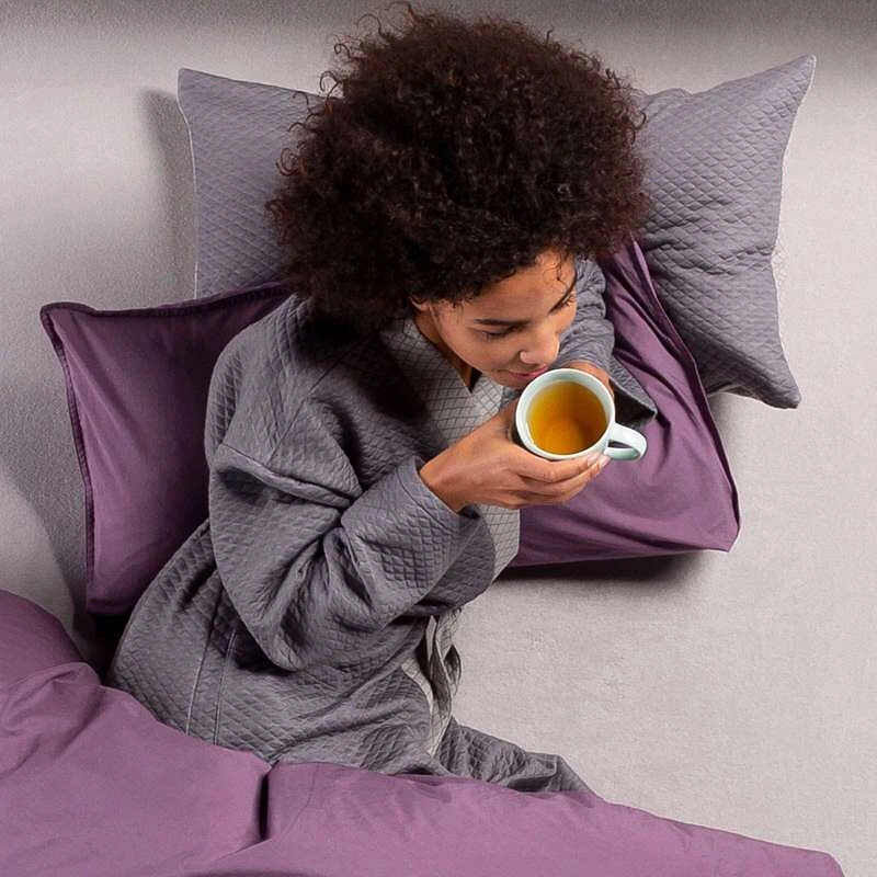 Frau in Hausmantel trinkt Tee im Bett, mit violetter Bettwäsche und Kissen