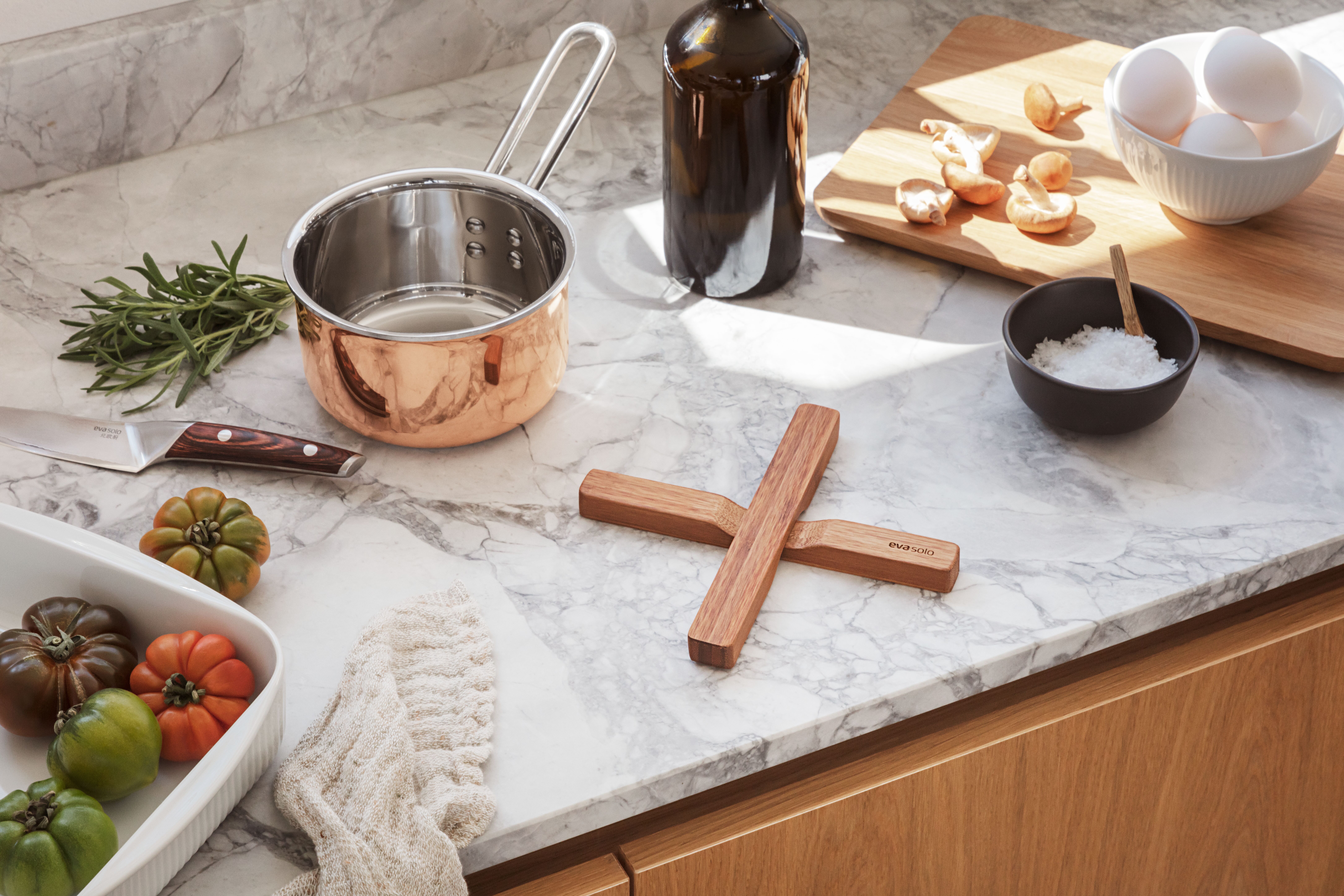 Accessoires von Eva Solo auf heller Küchenplatte mit Kupfertopf, Holztopfuntersetzer und Kräutern